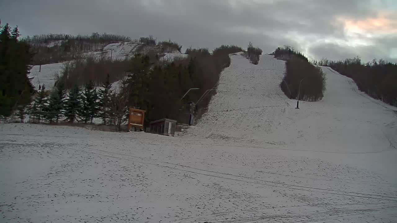 An overcast winter day reveals snow-covered ski slopes lined with evergreen and bare deciduous trees, featuring visible ski lift structures and small buildings at the base of the runs.