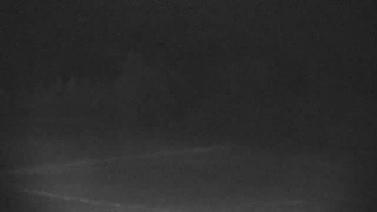 An extremely dark and grainy black-and-white image reveals an indistinct outdoor scene at night, with only faint variations in shade suggesting a distant horizon and vague landscape features.