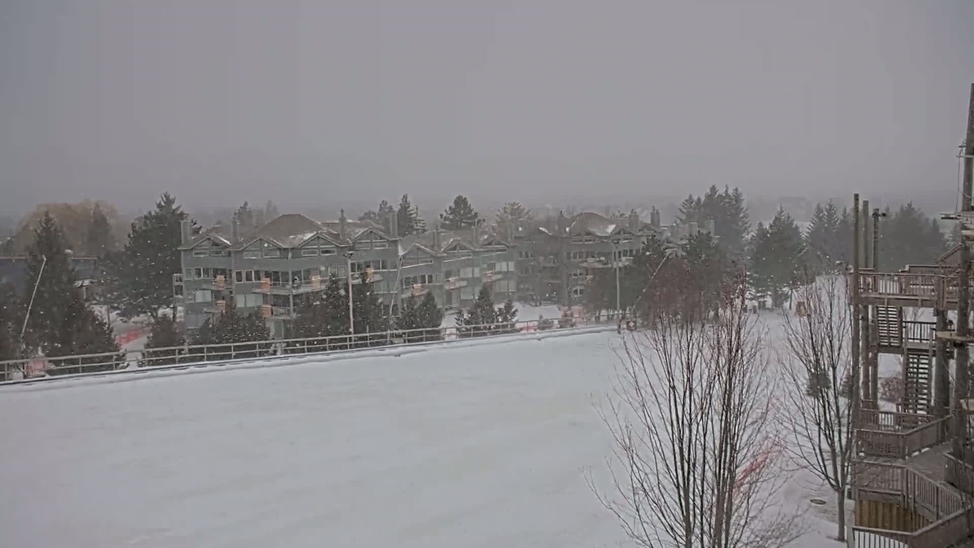 A snow-covered landscape features a row of multi-story buildings, bare trees, and a wooden structure, all under heavy snowfall.