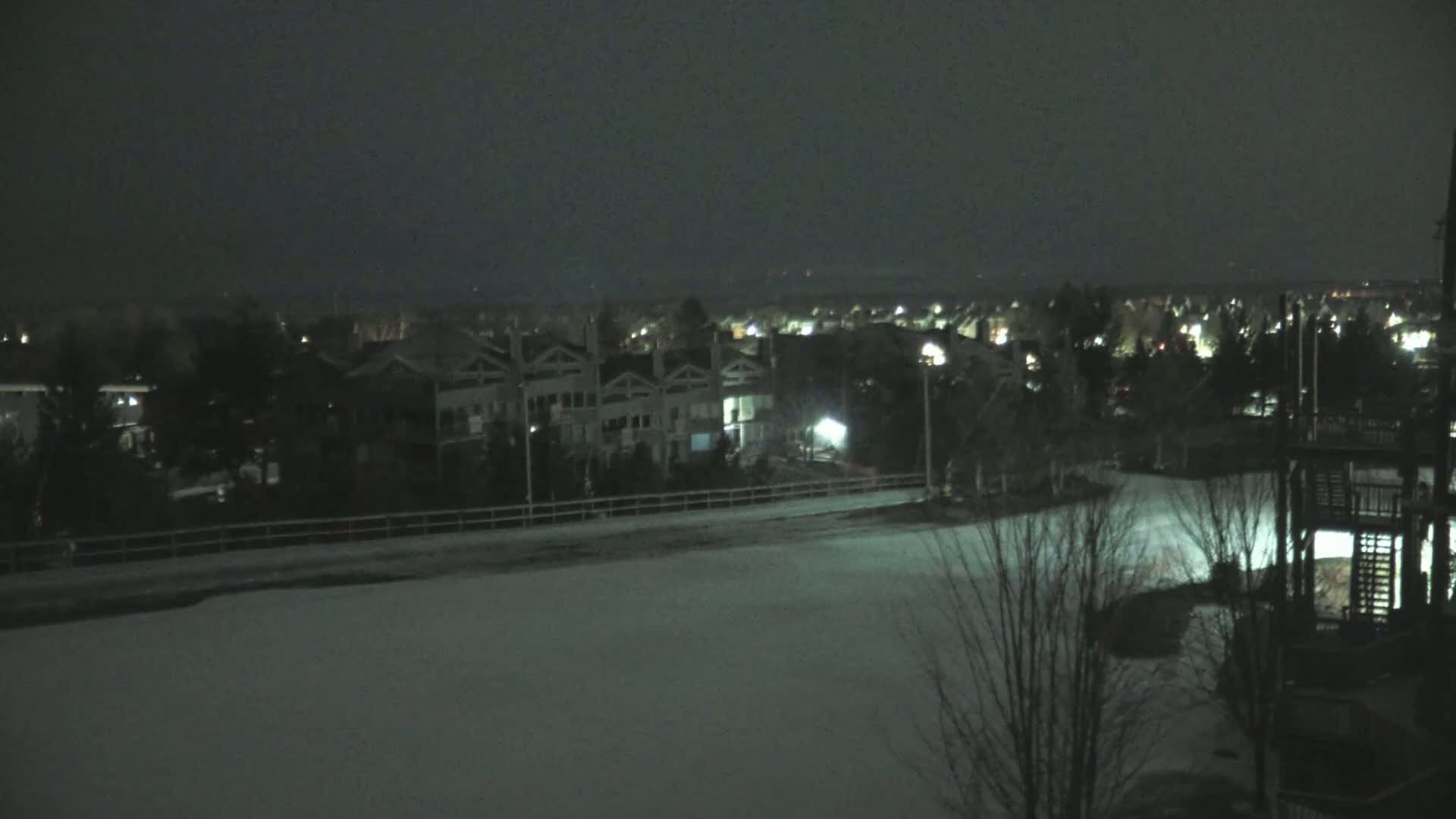 A snow-covered field at night is overlooked by a row of townhouses and a distant, dimly lit cityscape under a dark sky.