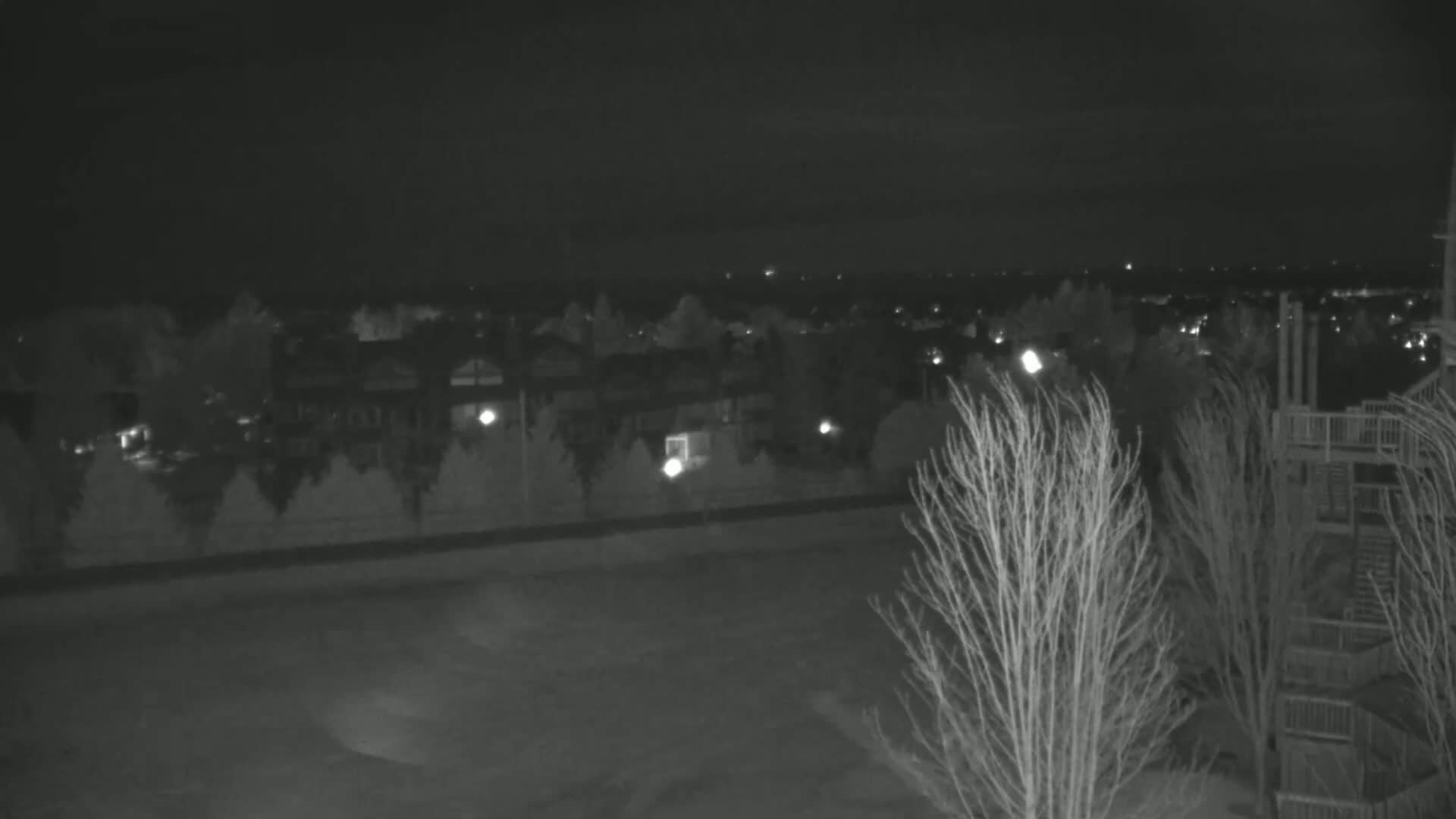 A nighttime view of a suburban area with a few lights on, some bare trees, and a section of a wooden structure in the bottom right corner.