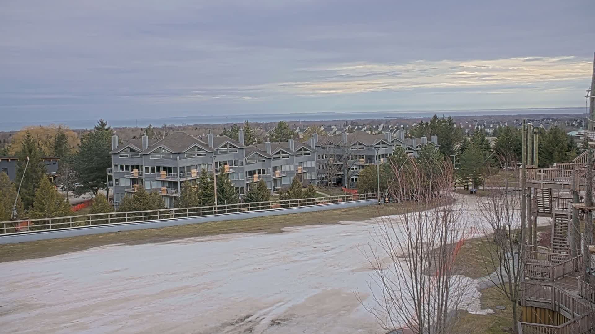 A row of gray multi-unit buildings sits on a hill overlooking a mostly snow-covered area and a calm body of water under a cloudy sky.