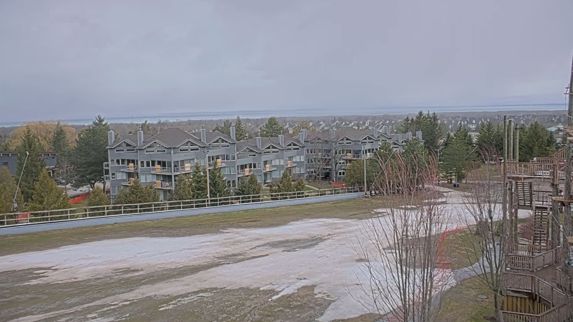 A row of gray apartment buildings sits near a mostly clear, snow-melting field overlooking a calm body of water under an overcast sky.