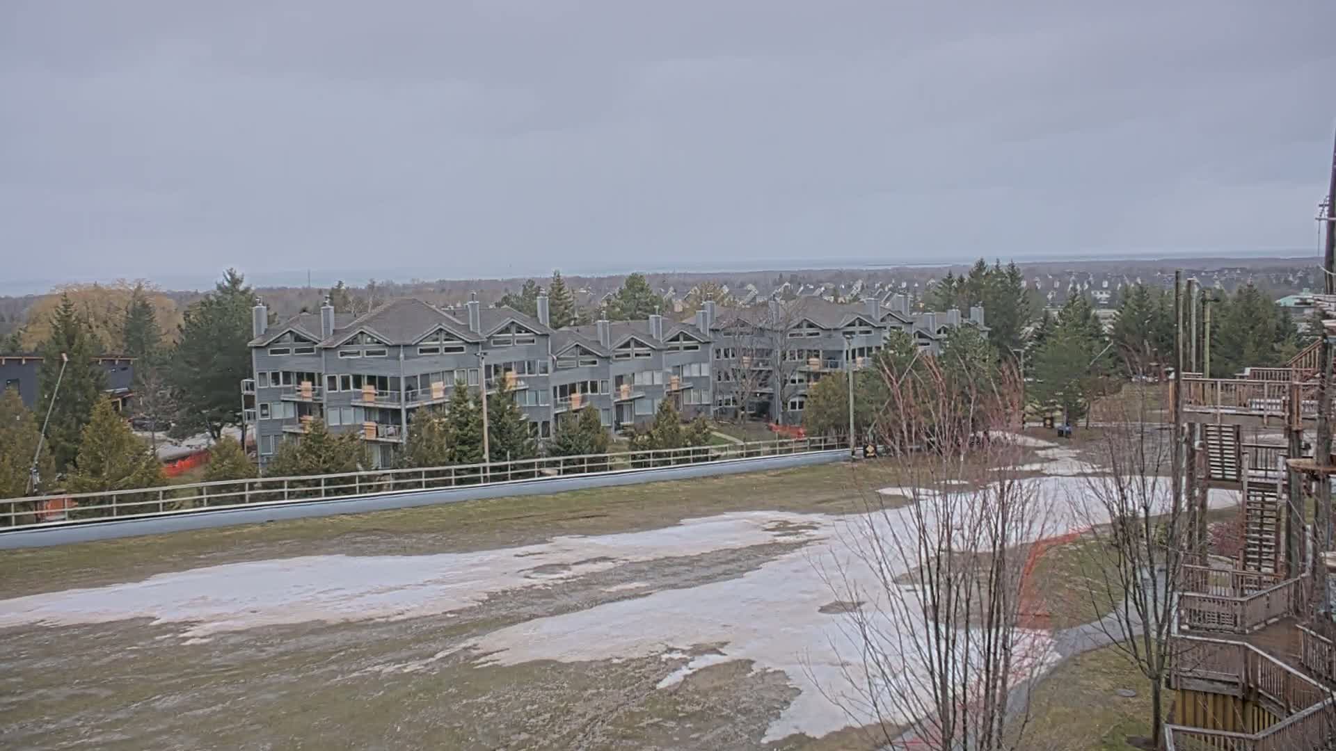 A row of gray multi-unit buildings sits on a hill overlooking a body of water under an overcast sky, with a partially snow-covered field in the foreground.