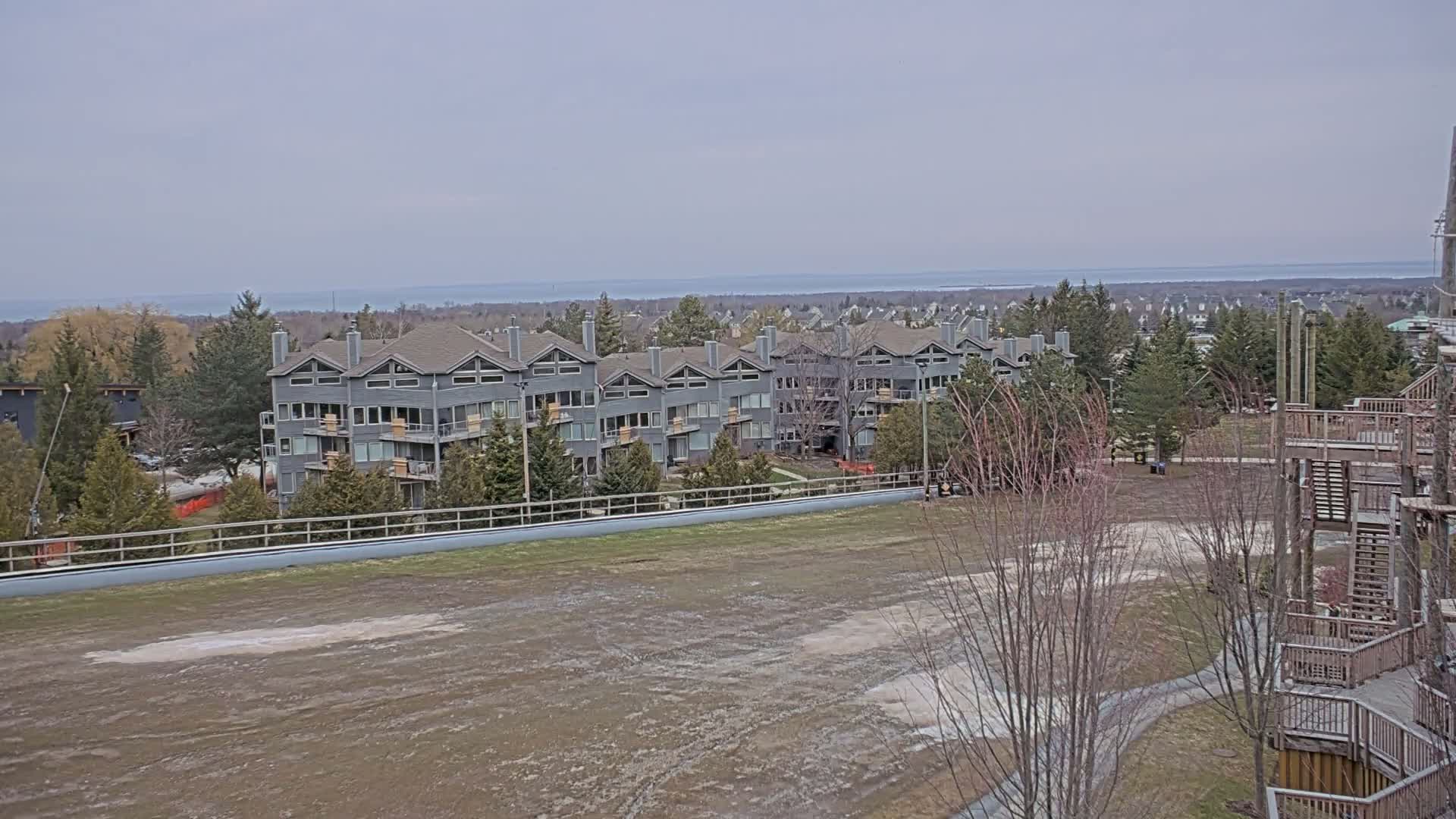 A long, gray apartment building sits behind a large, muddy, and sparsely vegetated field overlooking a calm body of water on an overcast day.