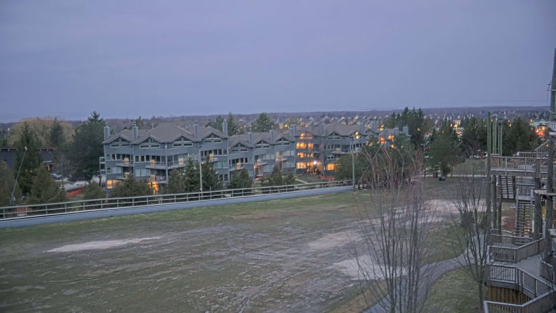 A row of gray multi-story buildings with lights on is situated beyond a large, barren, grassy field under a dusky sky.