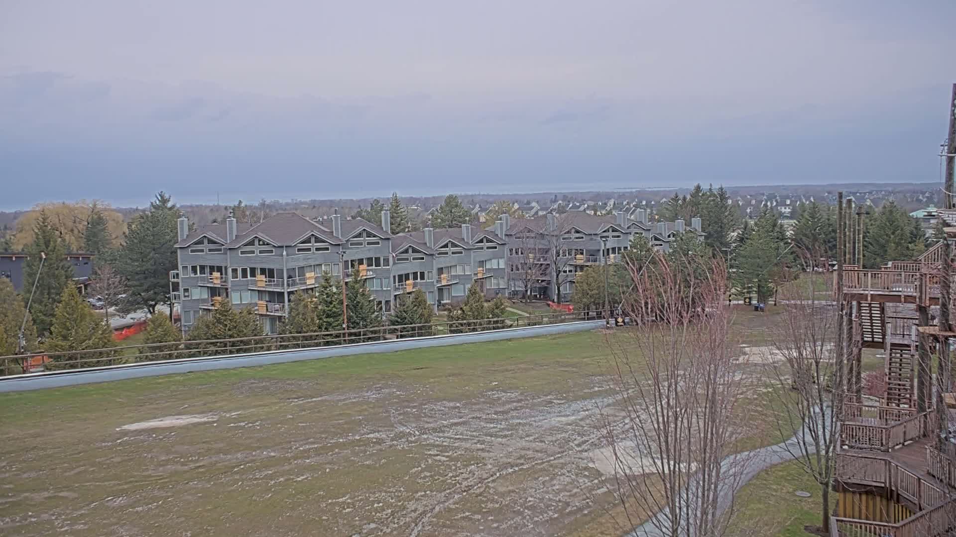 A row of gray multi-unit buildings sits on a hill overlooking a large, muddy field under an overcast sky.