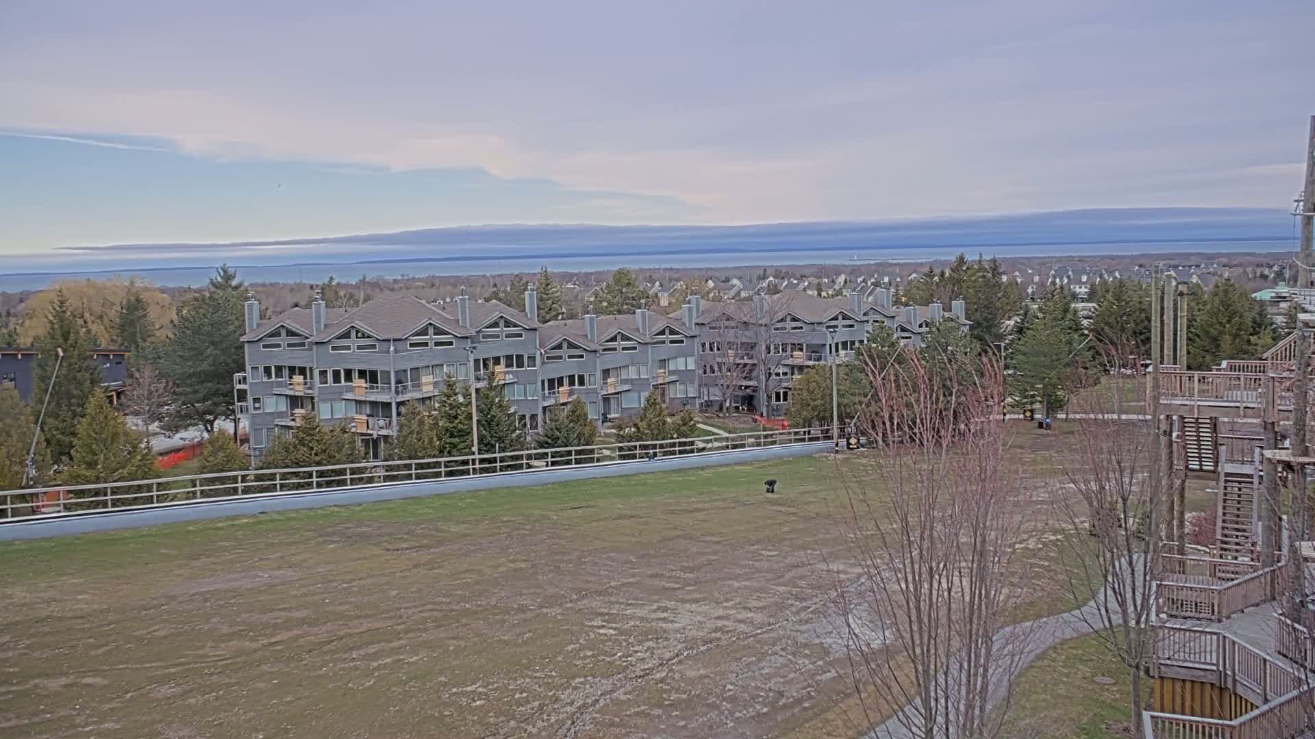 A gray multi-unit residential building overlooks a large grassy field and a calm body of water under a mostly cloudy sky.