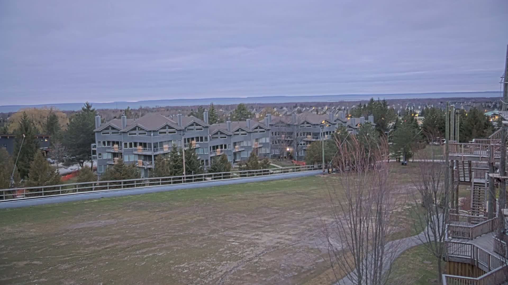 A long, gray apartment building sits behind a barren field overlooking a calm lake under a cloudy sky.