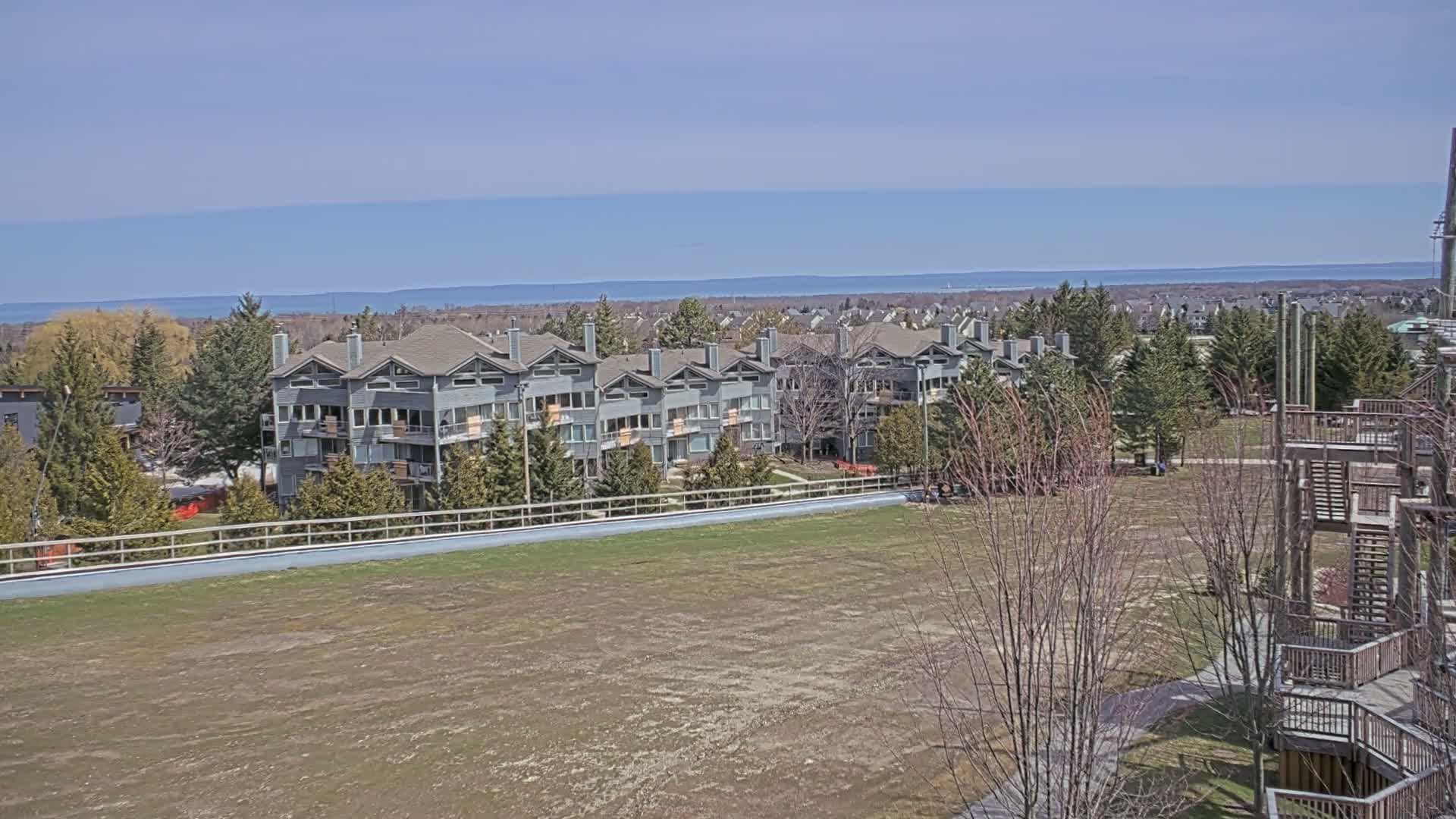 A row of gray multi-story buildings sits on a grassy hill overlooking a calm body of water under a clear sky.