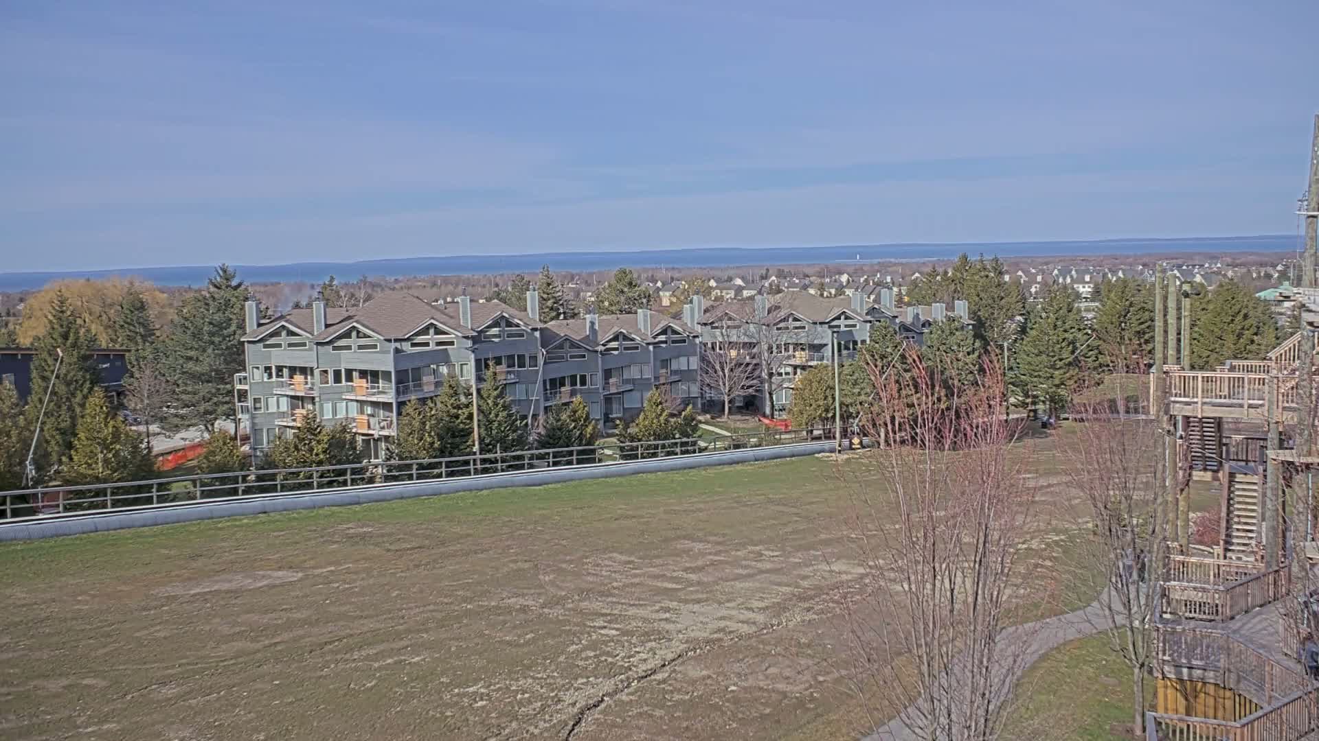 A row of gray multi-unit buildings sits on a grassy field overlooking a calm blue lake under a clear blue sky.