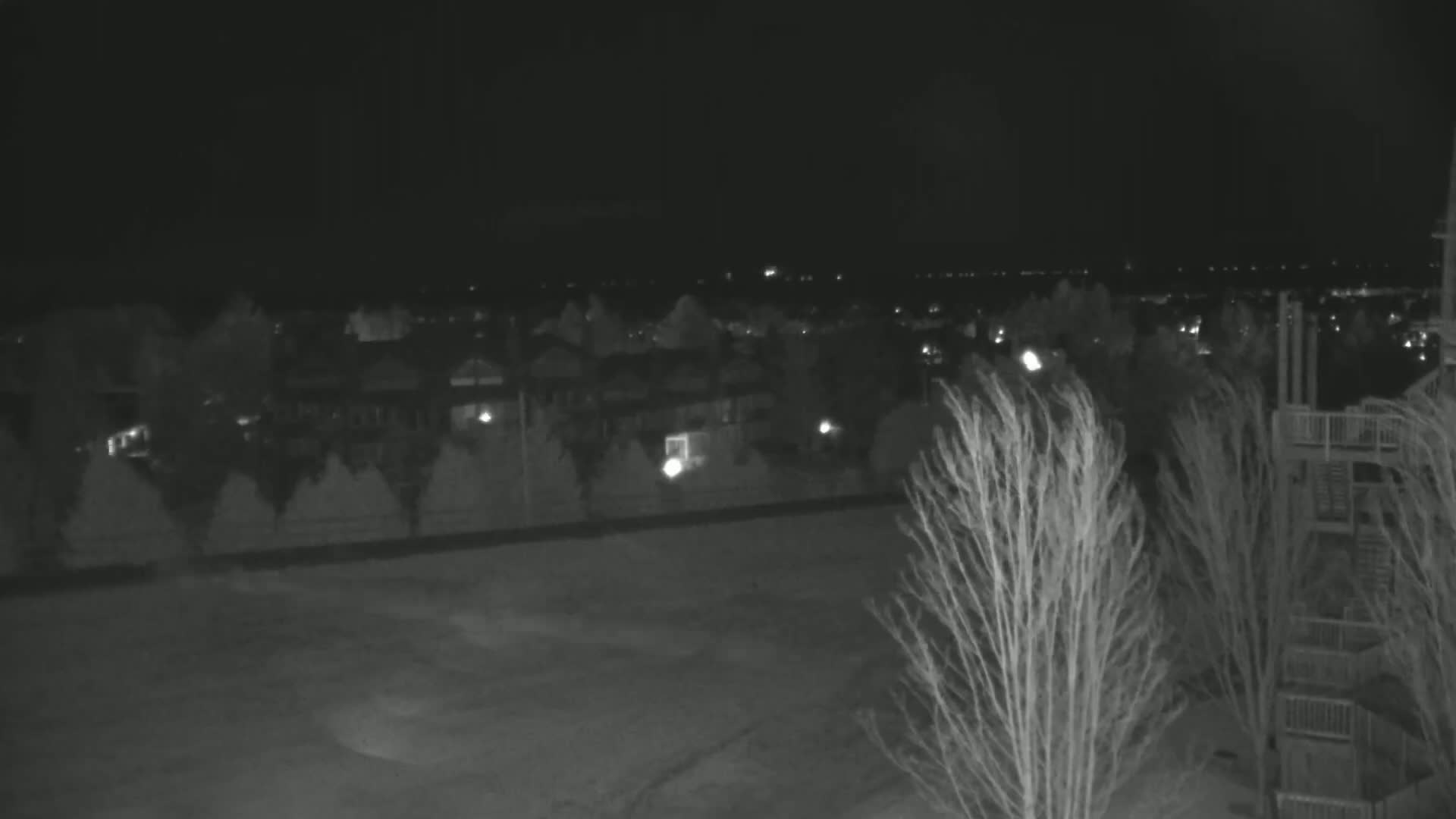 A nighttime, low-light view shows a residential area with sparsely lit houses and trees blowing in the wind.