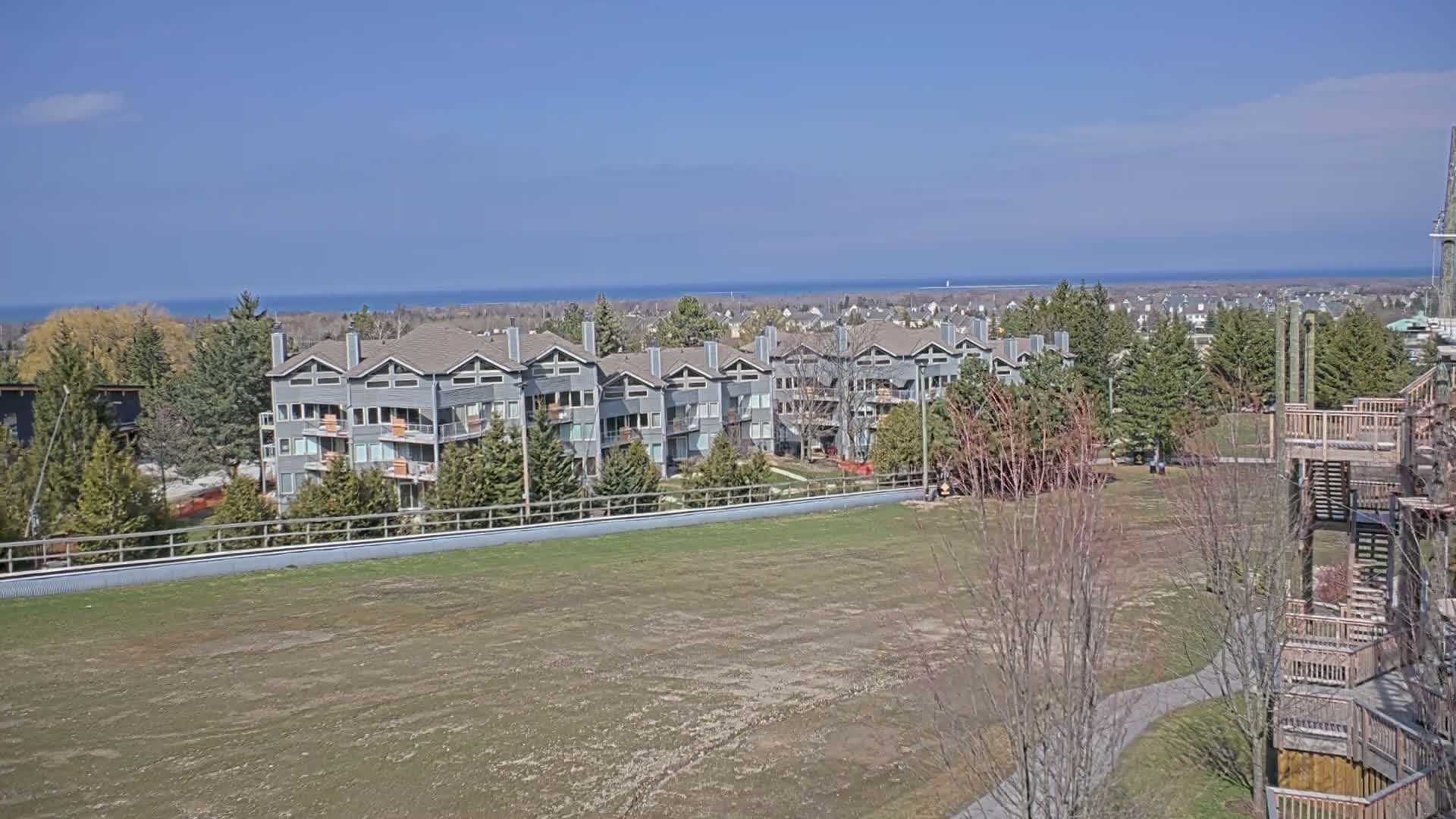 A row of gray apartment buildings sits behind a large, mostly bare, grassy field, overlooking a calm blue ocean on a clear, sunny day.