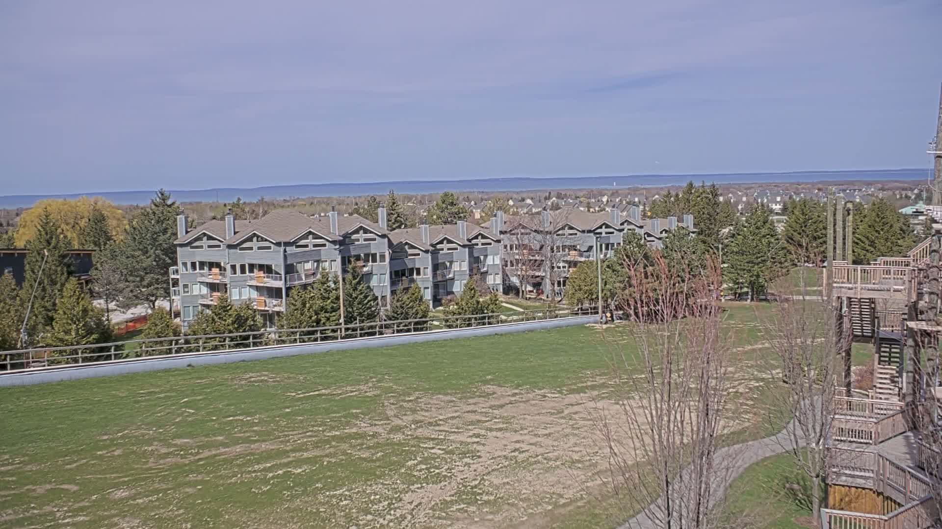 A row of gray multi-story buildings sits behind a large grassy field on a sunny day, with a body of water visible in the distance.