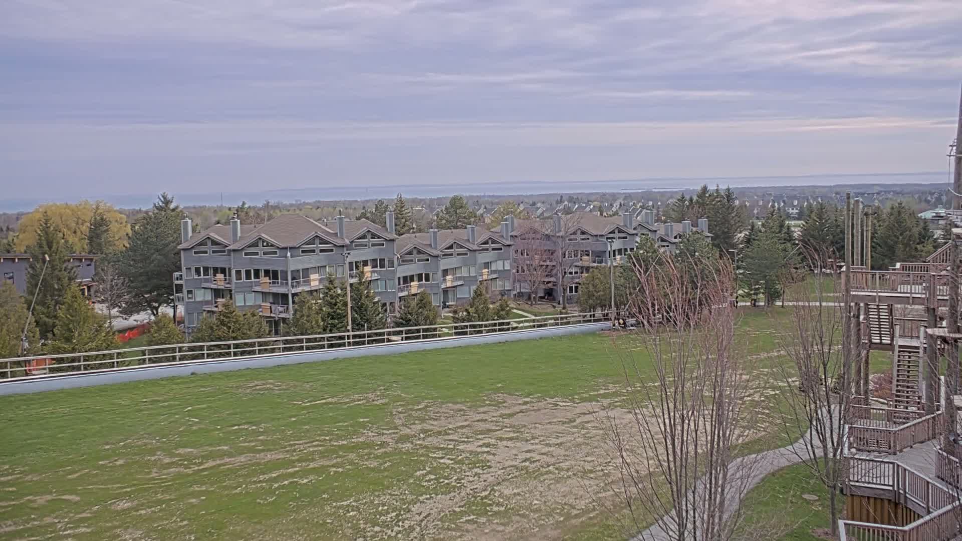 A gray multi-unit dwelling overlooks a patchy grassy field and a body of water under a cloudy sky.