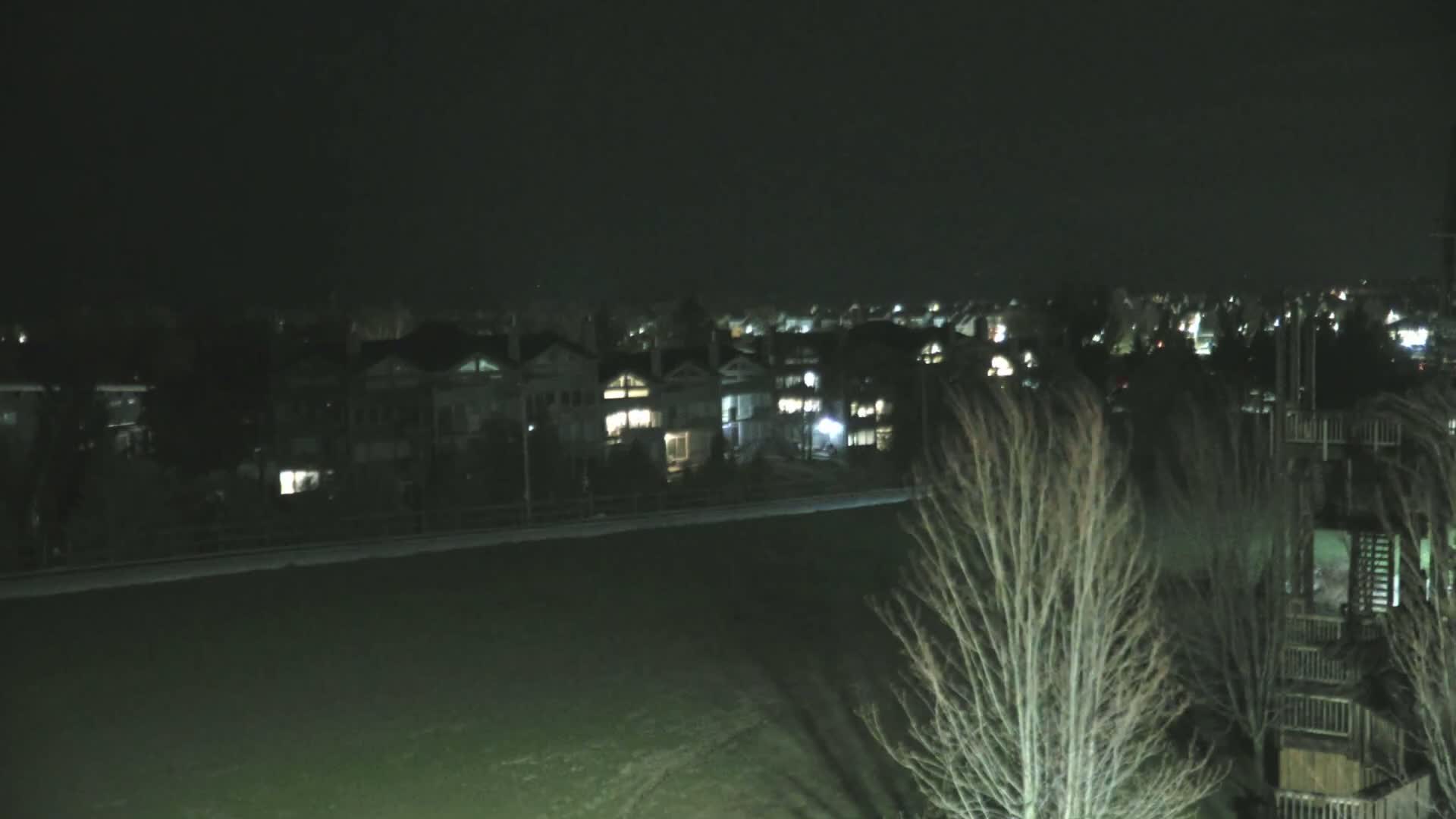 A dark, nighttime view shows a row of houses with illuminated windows across a grassy field, with bare trees in the foreground.