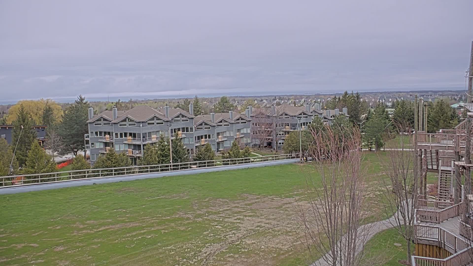 A gray multi-unit residential building is situated on a grassy field overlooking a distant body of water under an overcast sky.