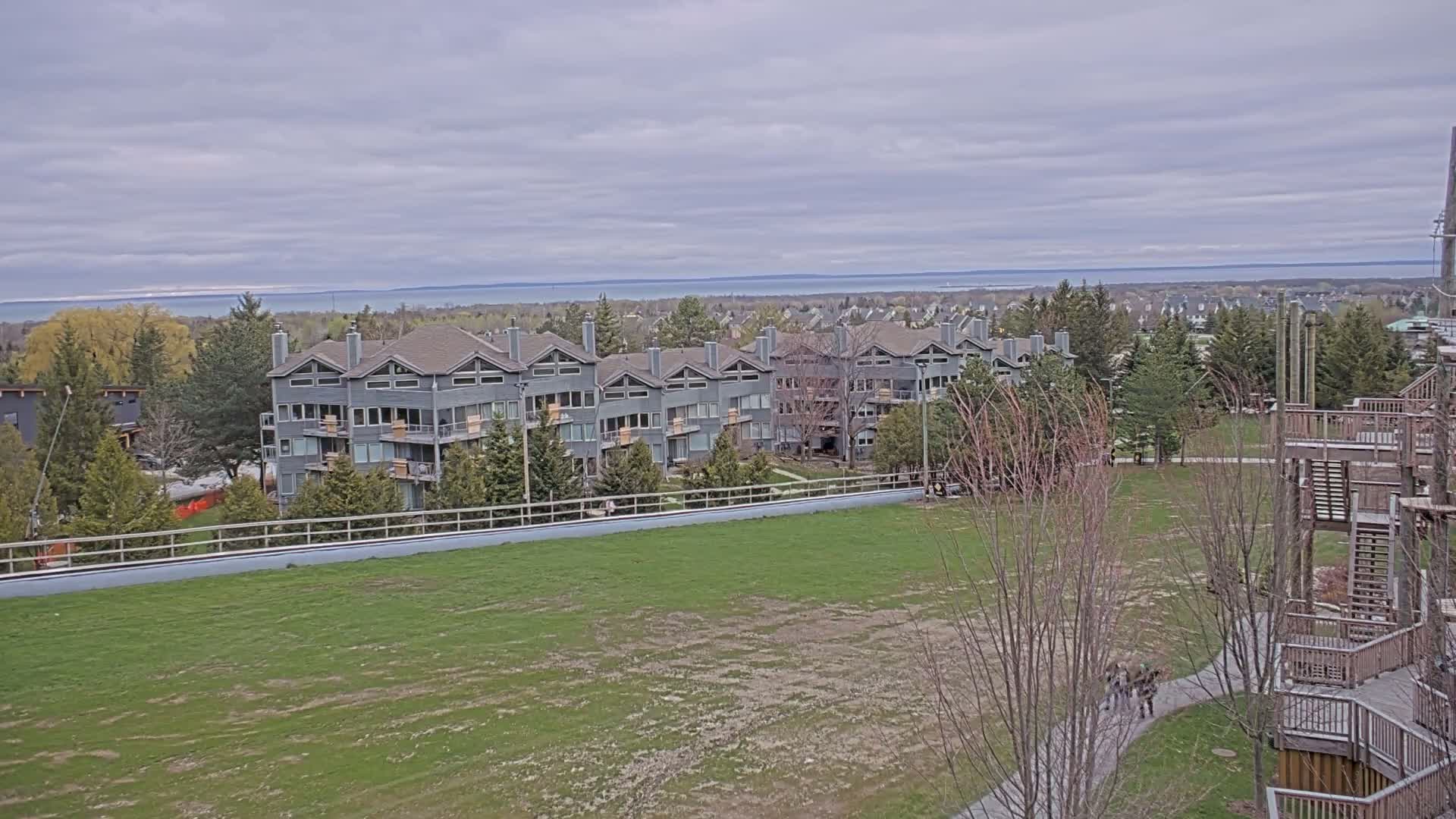 A row of gray multi-unit residential buildings sits on a grassy hill overlooking a body of water under a cloudy sky.
