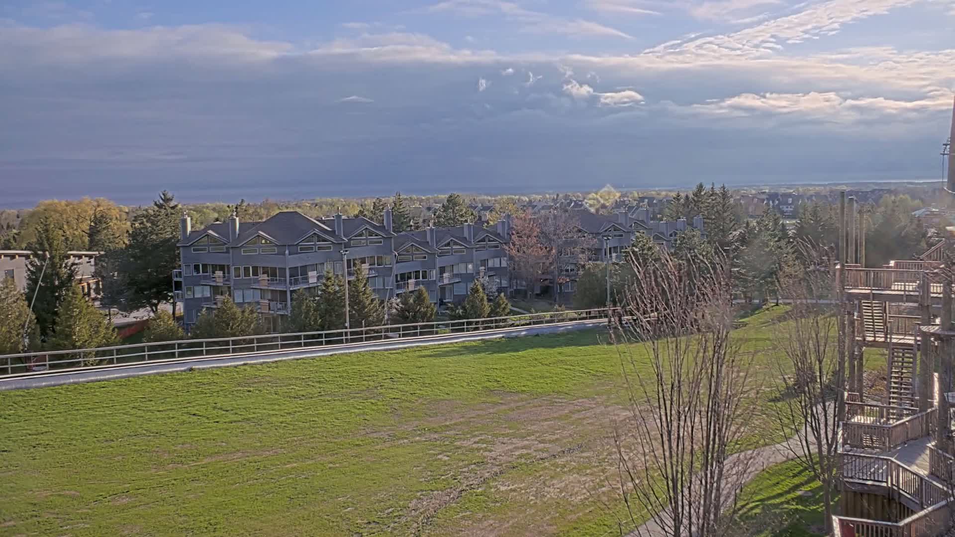 A row of gray multi-unit buildings sits beyond a grassy field under a partly cloudy sky.