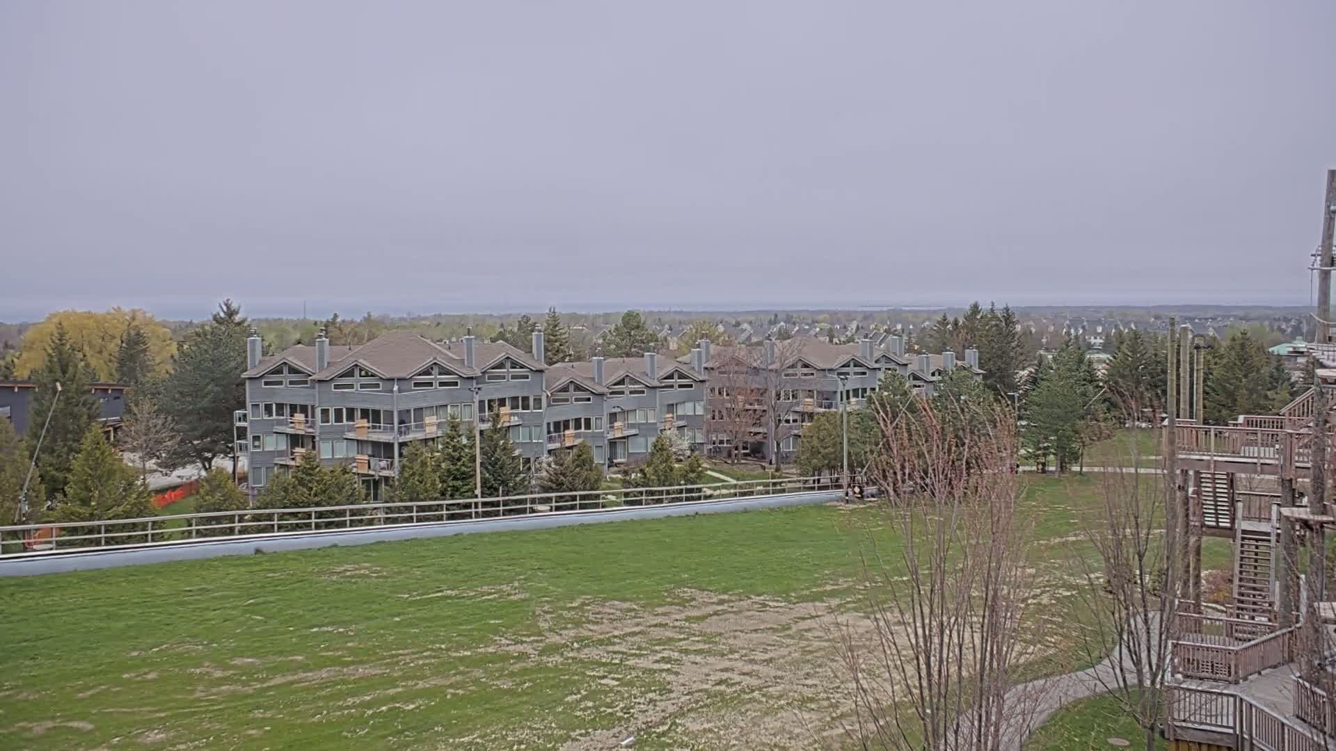 A row of gray multi-story buildings sits on a grassy hill overlooking a large expanse of green field and a distant body of water under an overcast sky.