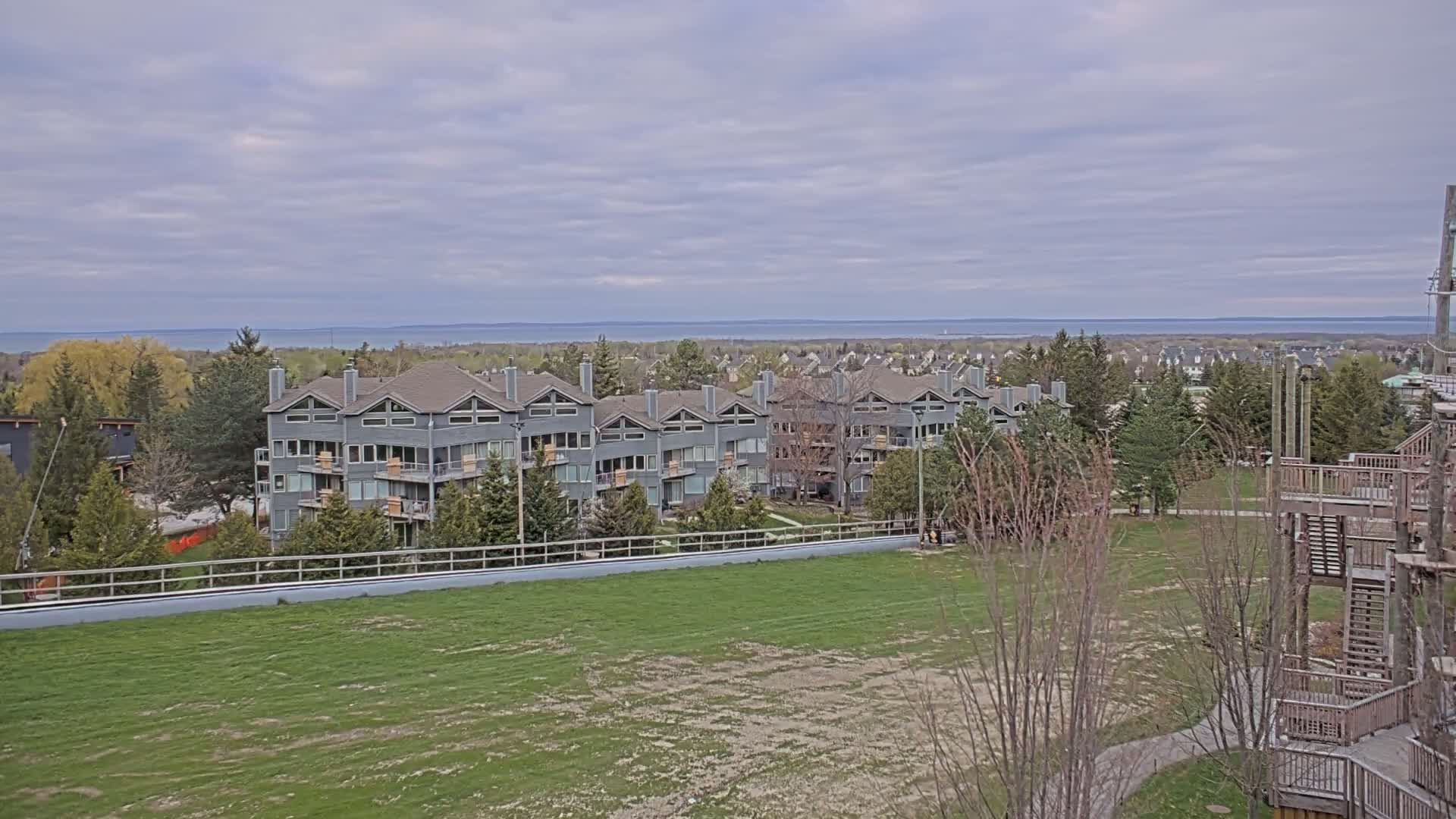 A row of gray multi-story buildings sits behind a grassy field overlooking a calm body of water under a cloudy sky.