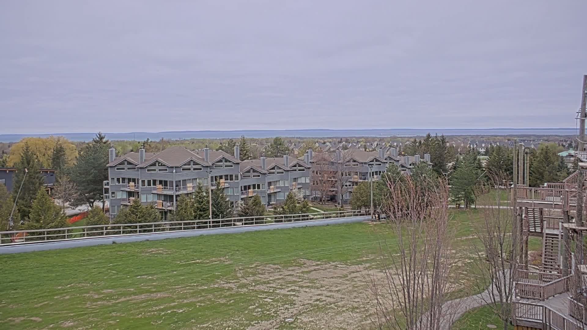 A gray multi-unit residential building sits on a grassy hill overlooking a calm body of water under an overcast sky.