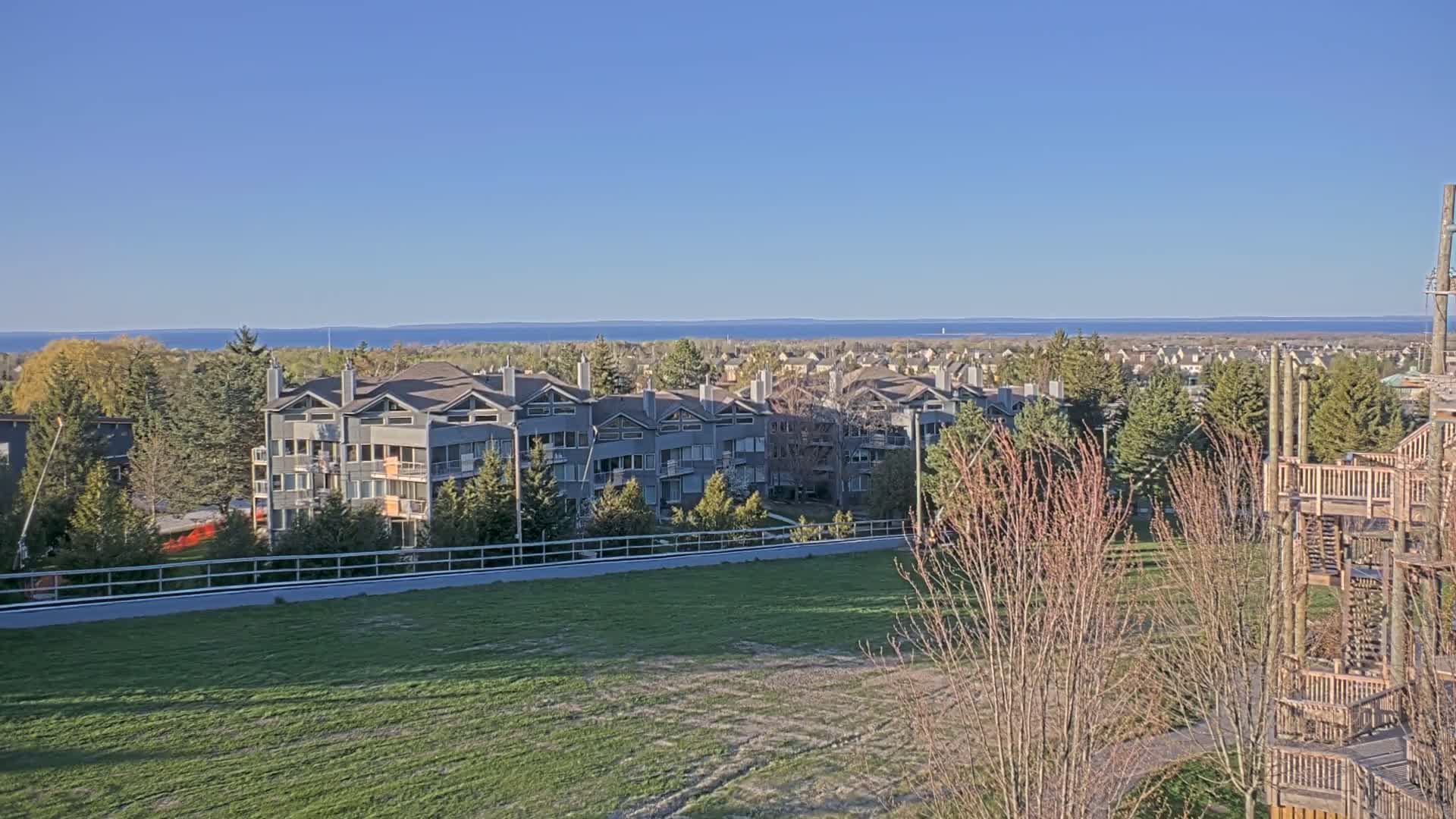 A row of gray multi-unit buildings sits on a grassy area overlooking a calm blue body of water under a clear blue sky.