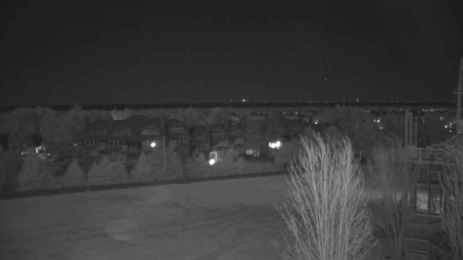 A nighttime view shows a residential area with sparsely lit houses, trees, and a distant horizon under a clear, dark sky.