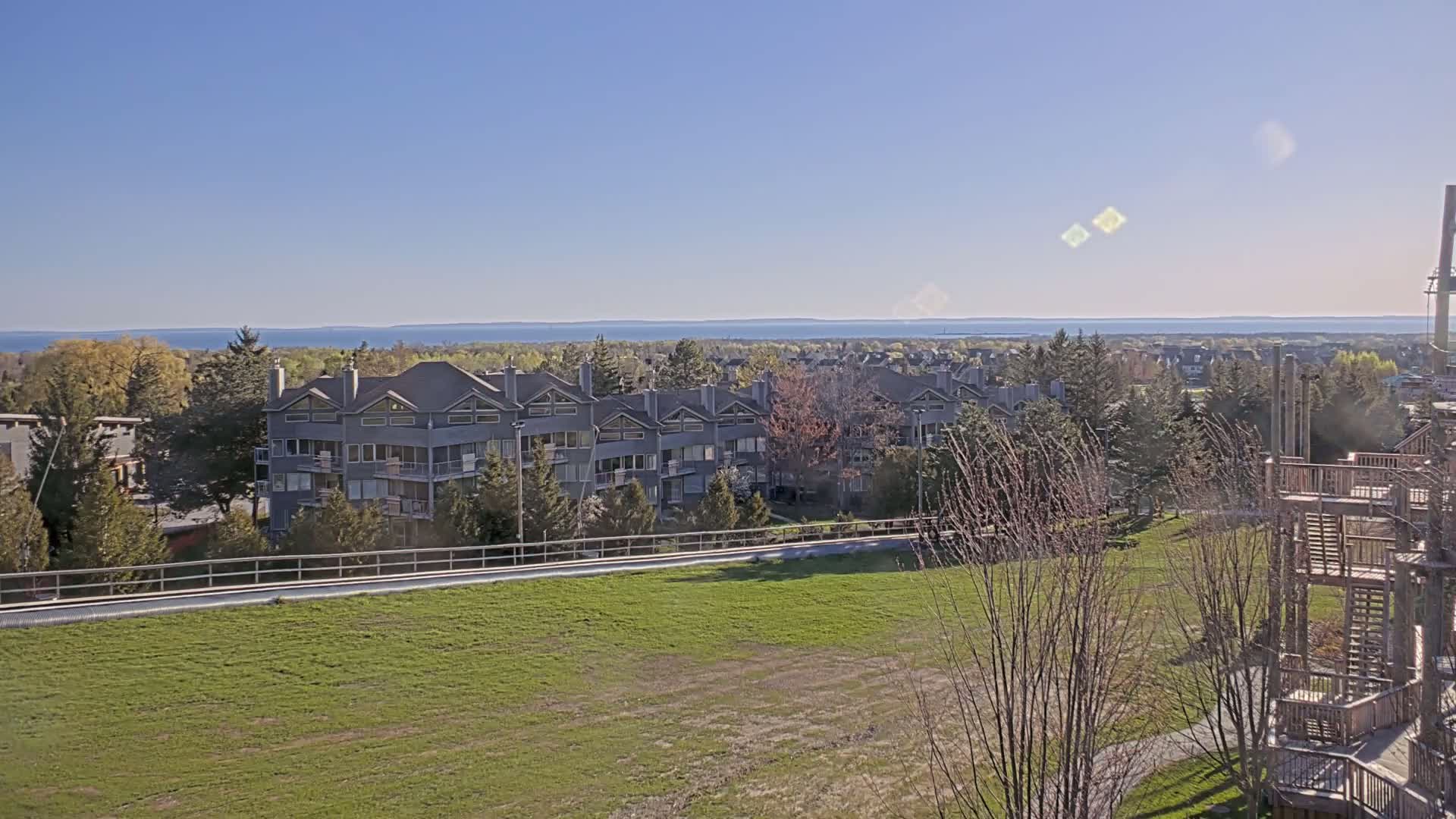 A row of gray townhouses sits on a grassy hill overlooking a calm blue lake under a clear blue sky.