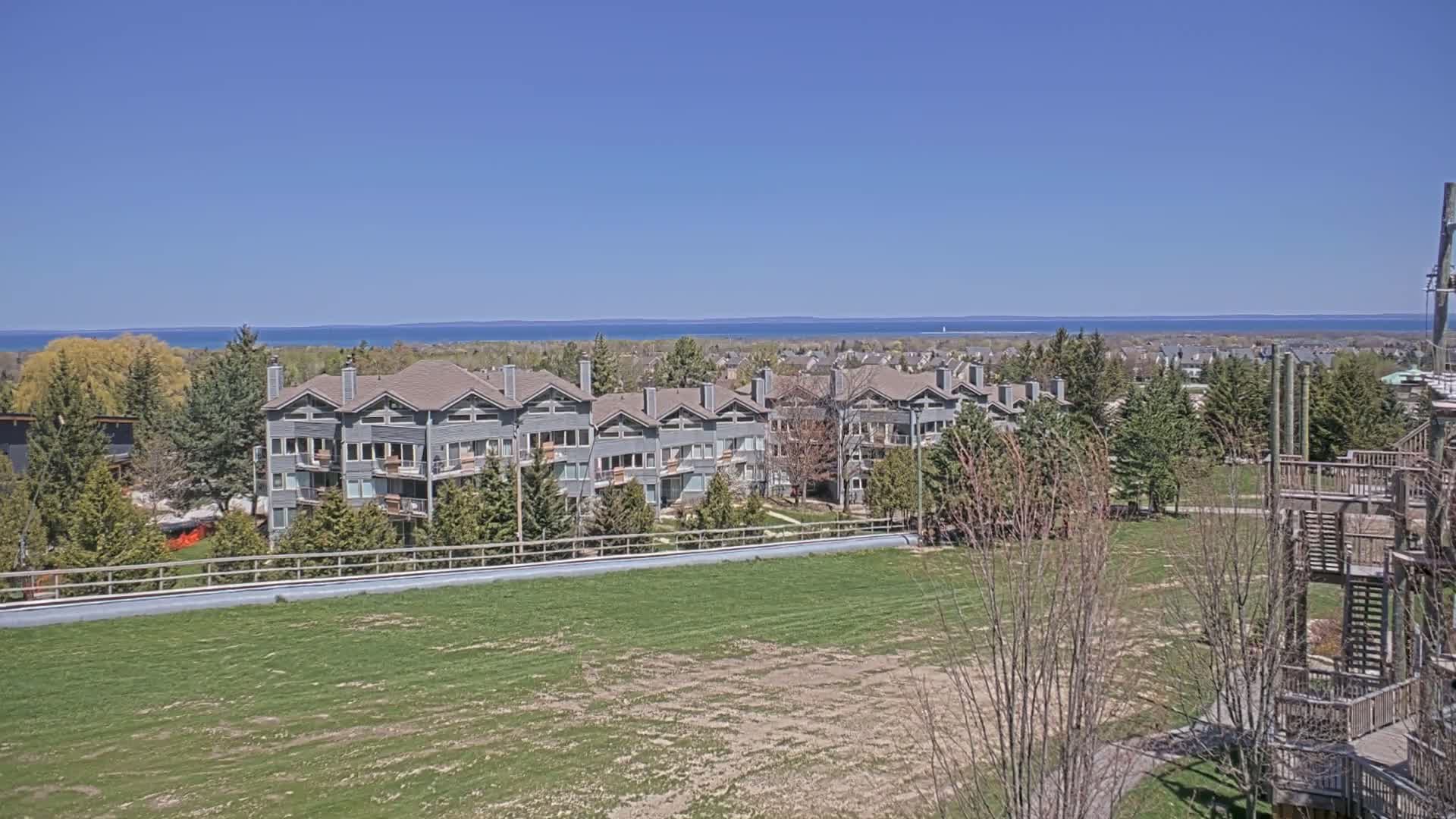 A row of gray multi-unit buildings overlooks a grassy field and a calm blue lake under a clear blue sky.