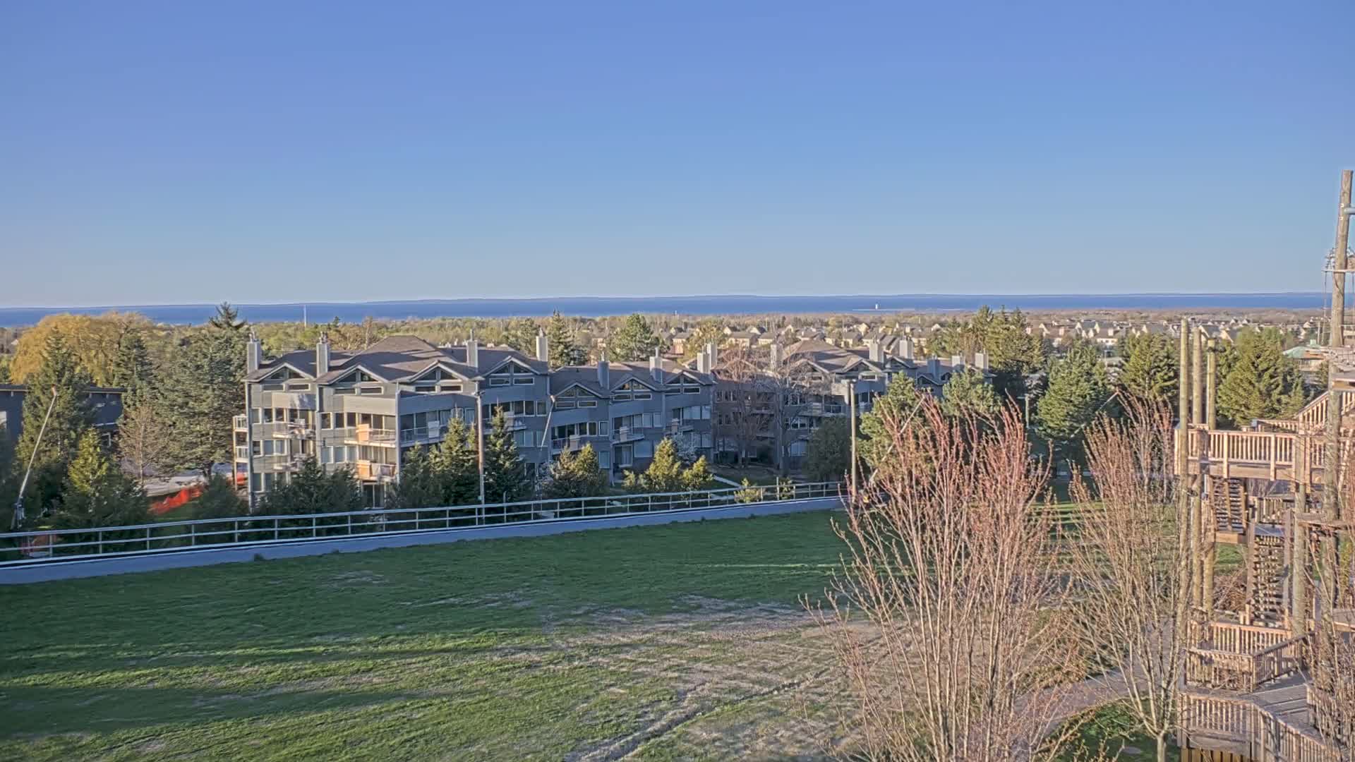 A row of gray multi-story buildings sits atop a grassy hill overlooking a calm blue lake under a clear blue sky.