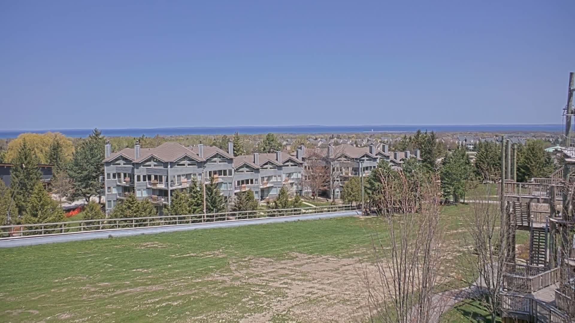 A row of gray multi-story buildings sits on a grassy hill overlooking a calm blue lake under a clear blue sky.