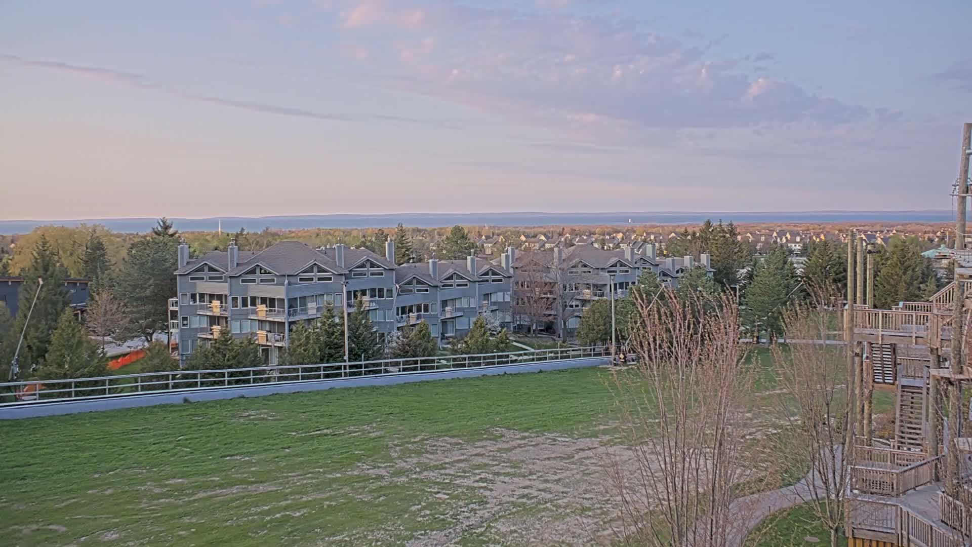 A row of gray townhouses overlooks a grassy field, a body of water, and a distant residential area under a partly cloudy, sunset sky.