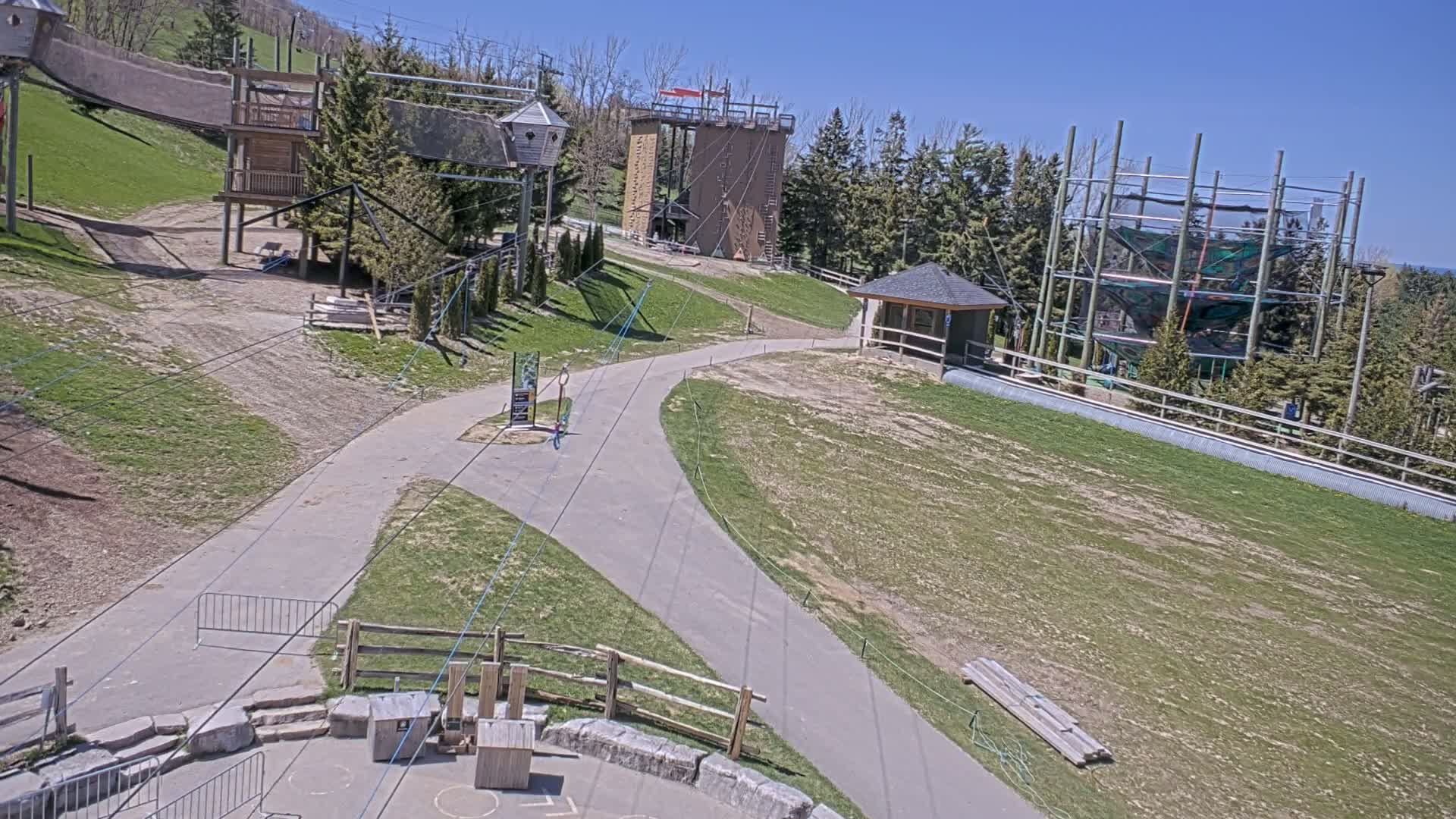A paved pathway winds through a grassy area featuring various outdoor recreational structures, including climbing towers and zip line equipment, under a clear blue sky.