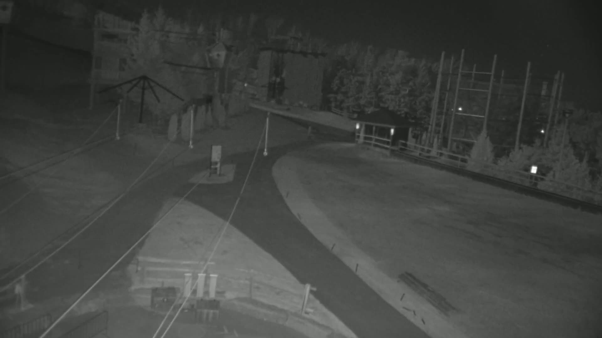 A nighttime, low-light view shows an empty paved area with intersecting paths, surrounded by buildings and trees.