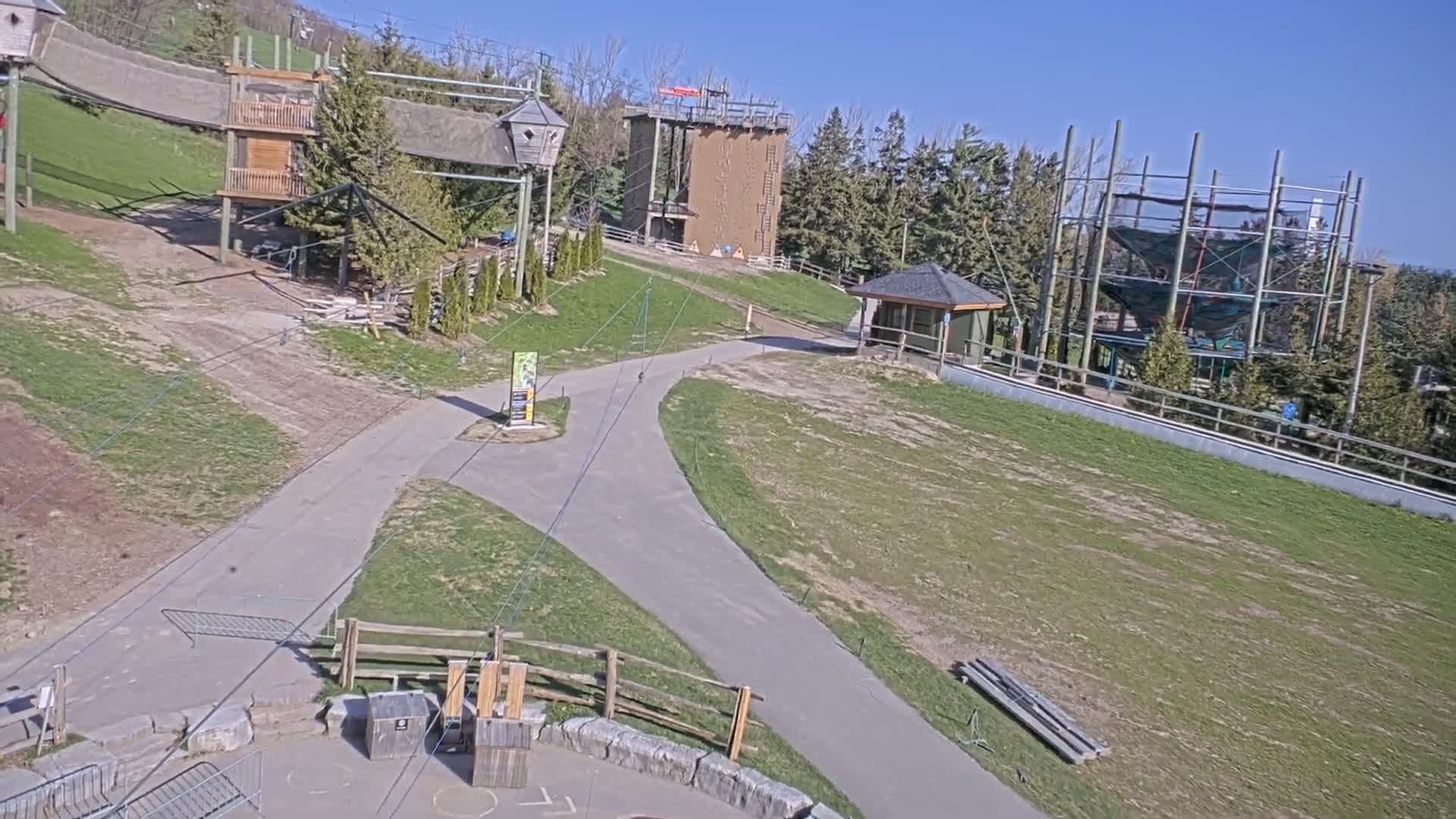 A paved pathway winds through a grassy outdoor area with various wooden structures, including a climbing wall and a large net structure, under a bright, sunny sky.