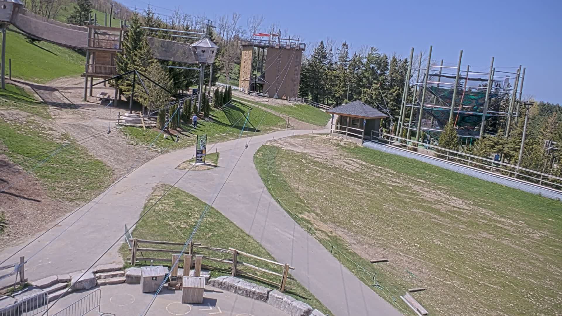 A paved path winds through a grassy area with various climbing structures and zip lines, under a clear blue sky.