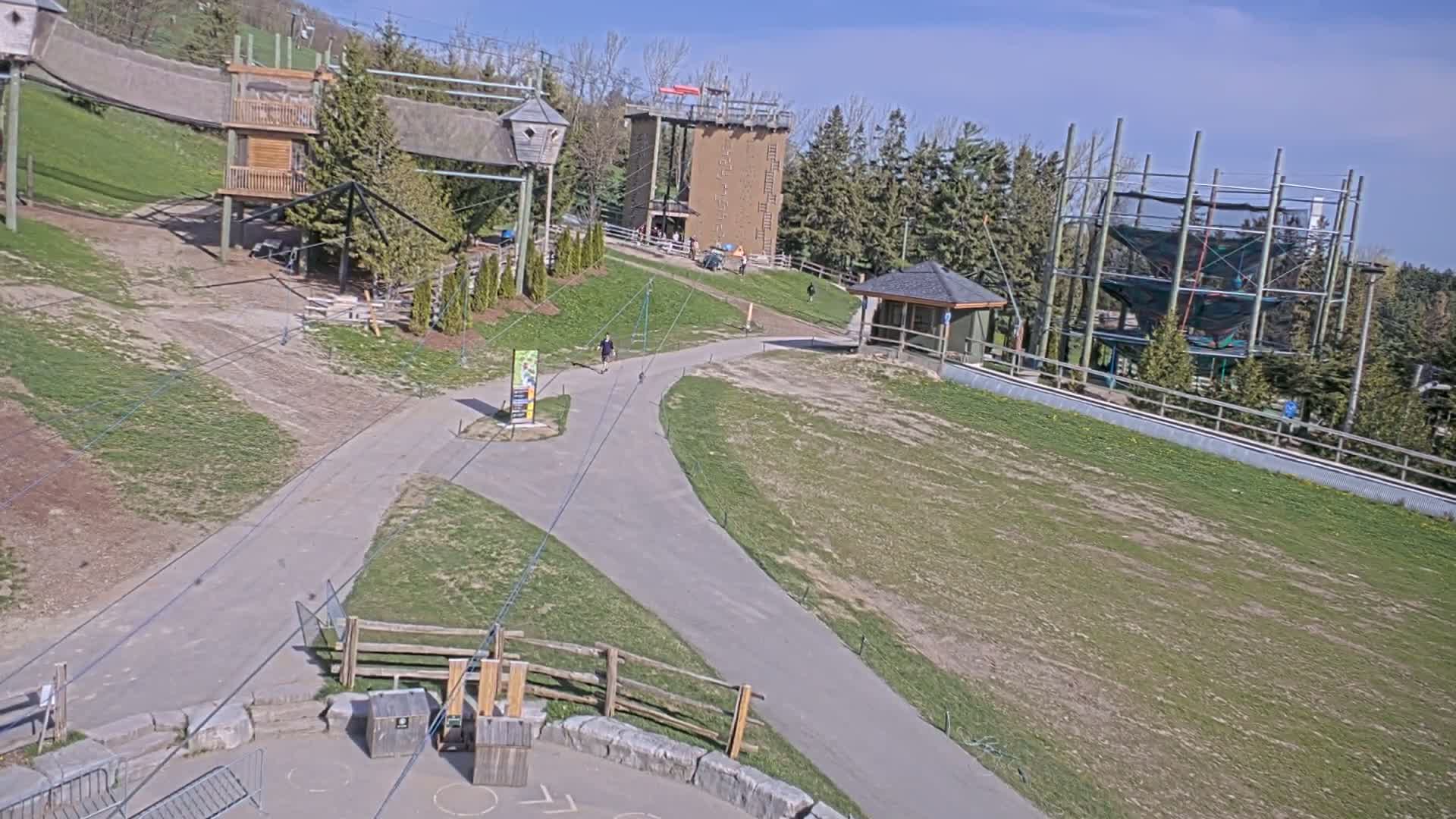 An outdoor scene on a sunny day shows a paved pathway winding through a grassy area with various wooden structures, including elevated platforms and a climbing tower.