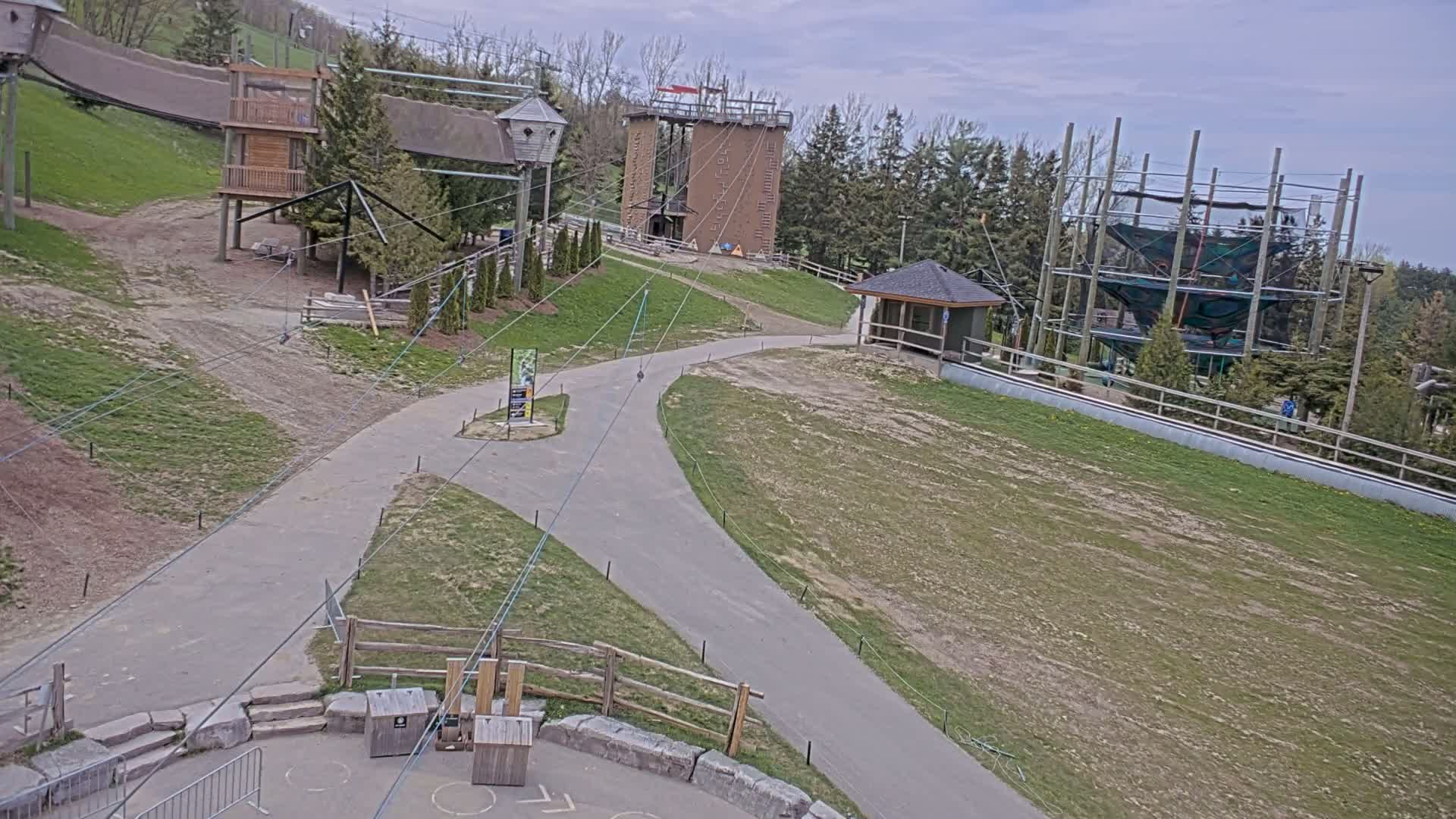An outdoor scene on a partly cloudy day shows paved paths winding through a grassy area with various climbing structures and a zip line course.