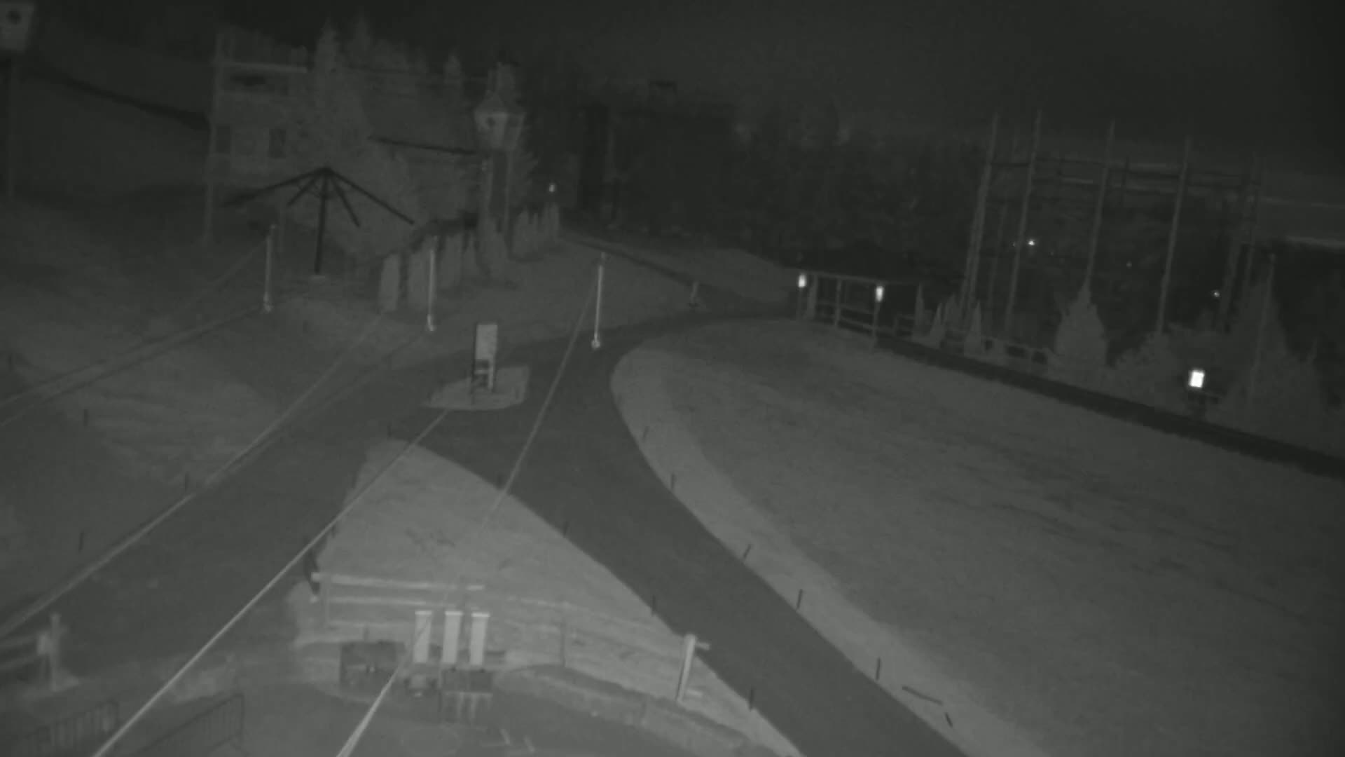 A nighttime, low-light view of a snow-covered intersection with a building and a structure resembling a tall fence in the background.