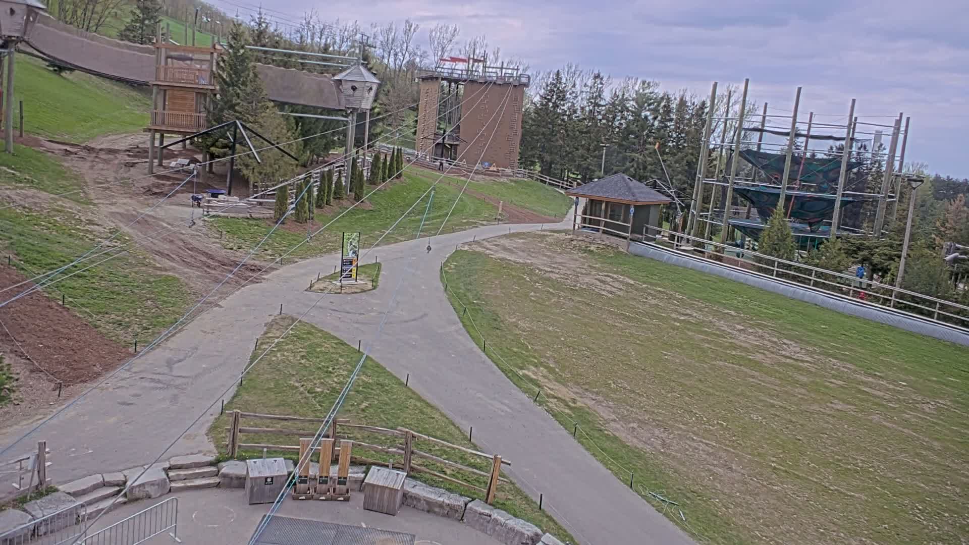 An outdoor area with paved paths, grassy fields, and several structures including climbing towers and treetop adventure courses, under a partly cloudy sky.