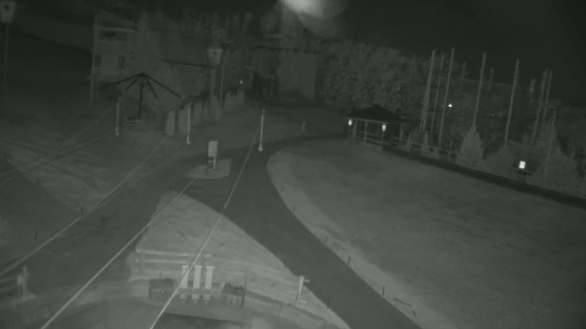 A nighttime, overhead view shows a paved road curving through a sparsely vegetated area near buildings and tall poles, under a slightly hazy sky.