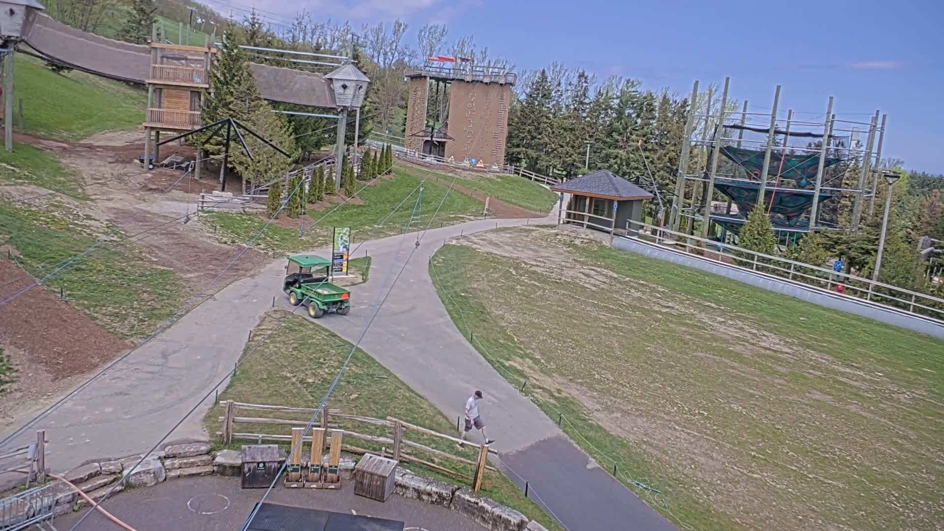 A person walks on a paved path near a green utility vehicle, climbing structures, and a ropes course on a grassy hillside under a mostly clear sky.