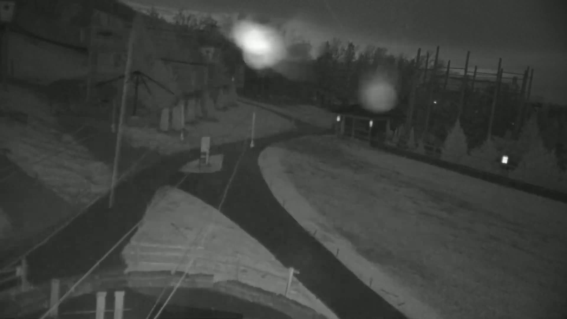 A nighttime, snow-covered scene shows a paved path curving through a residential area near some trees and buildings under a dark, cloudy sky.