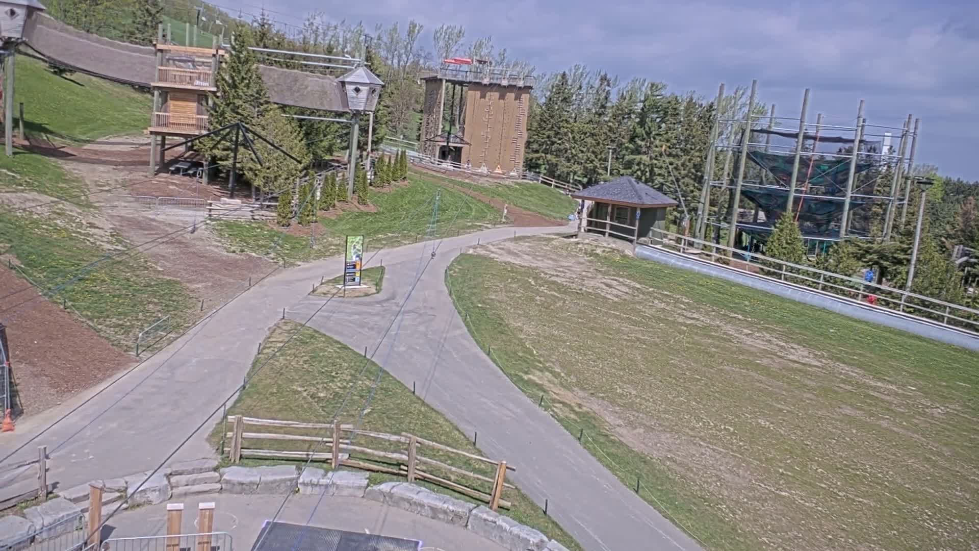 An outdoor recreational area with paved paths, various wooden structures, a climbing wall, and a tall netted climbing tower under a partly sunny sky.