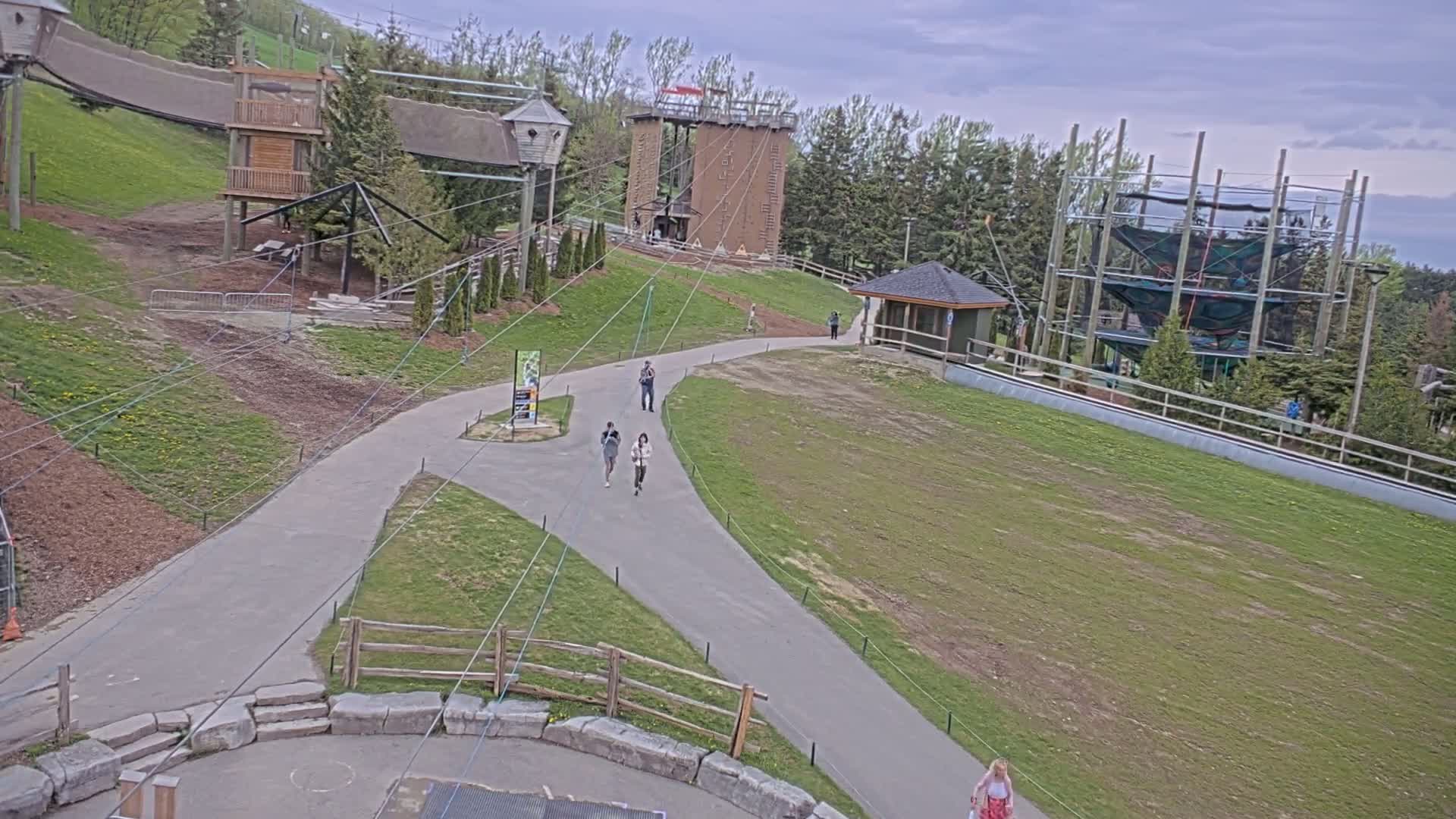 A paved pathway winds through a grassy area with several people walking, leading to various wooden structures, including a climbing tower and a ropes course, under partly cloudy skies.