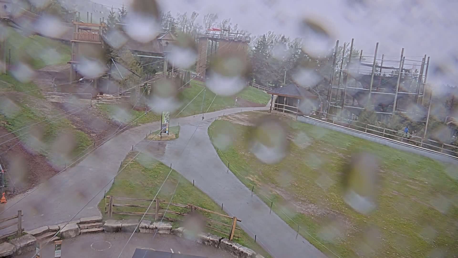 A rainy view of paved pathways winding through a grassy area with various structures, including a climbing tower and a gazebo, partially obscured by rain droplets on a window.