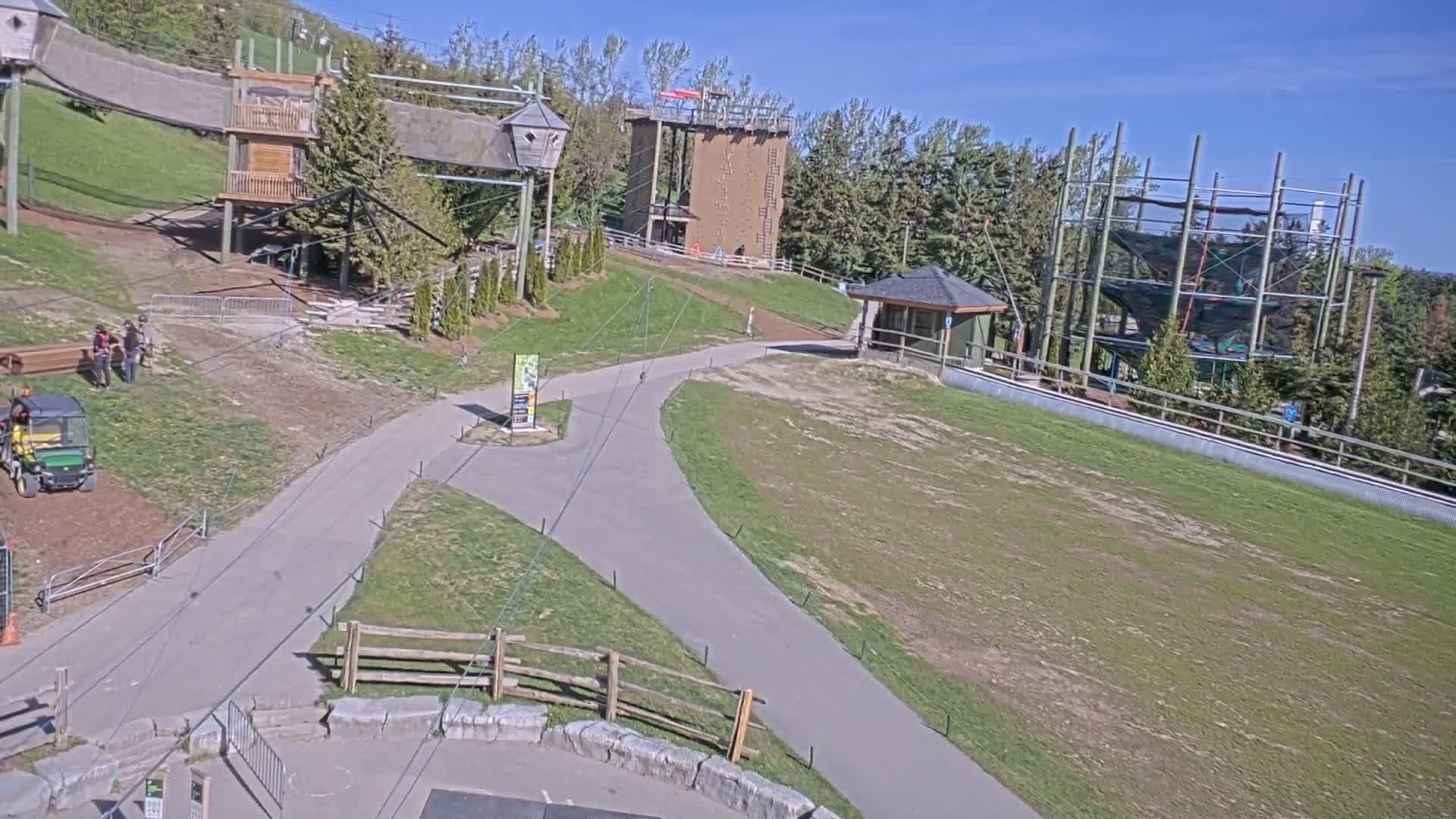A paved pathway winds through a grassy area with various outdoor recreational structures, including climbing walls and a ropes course, under a sunny sky.
