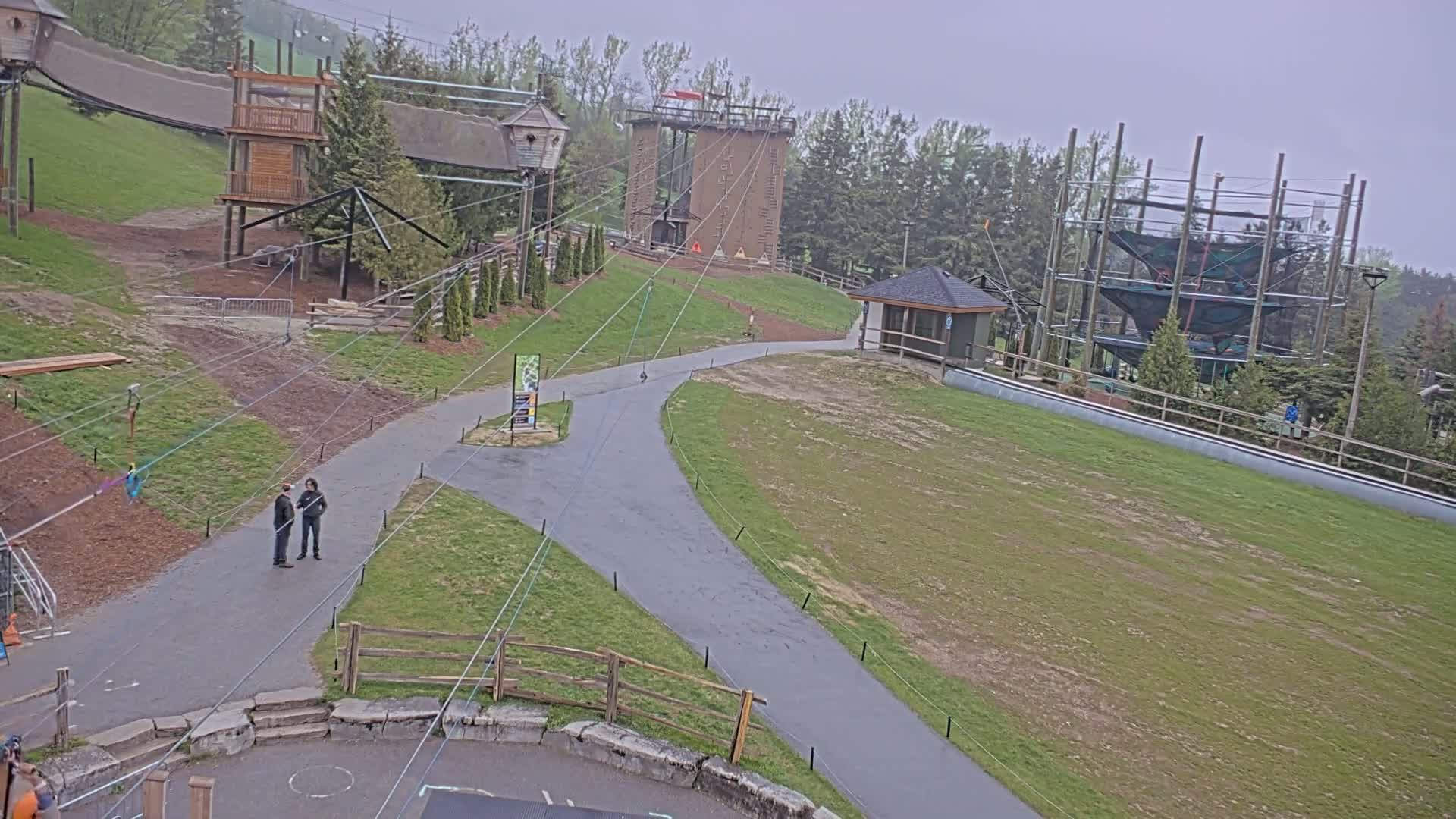 An outdoor scene on an overcast day shows a paved pathway winding through grassy areas past several wooden structures, including a climbing tower and a multi-level rope course.