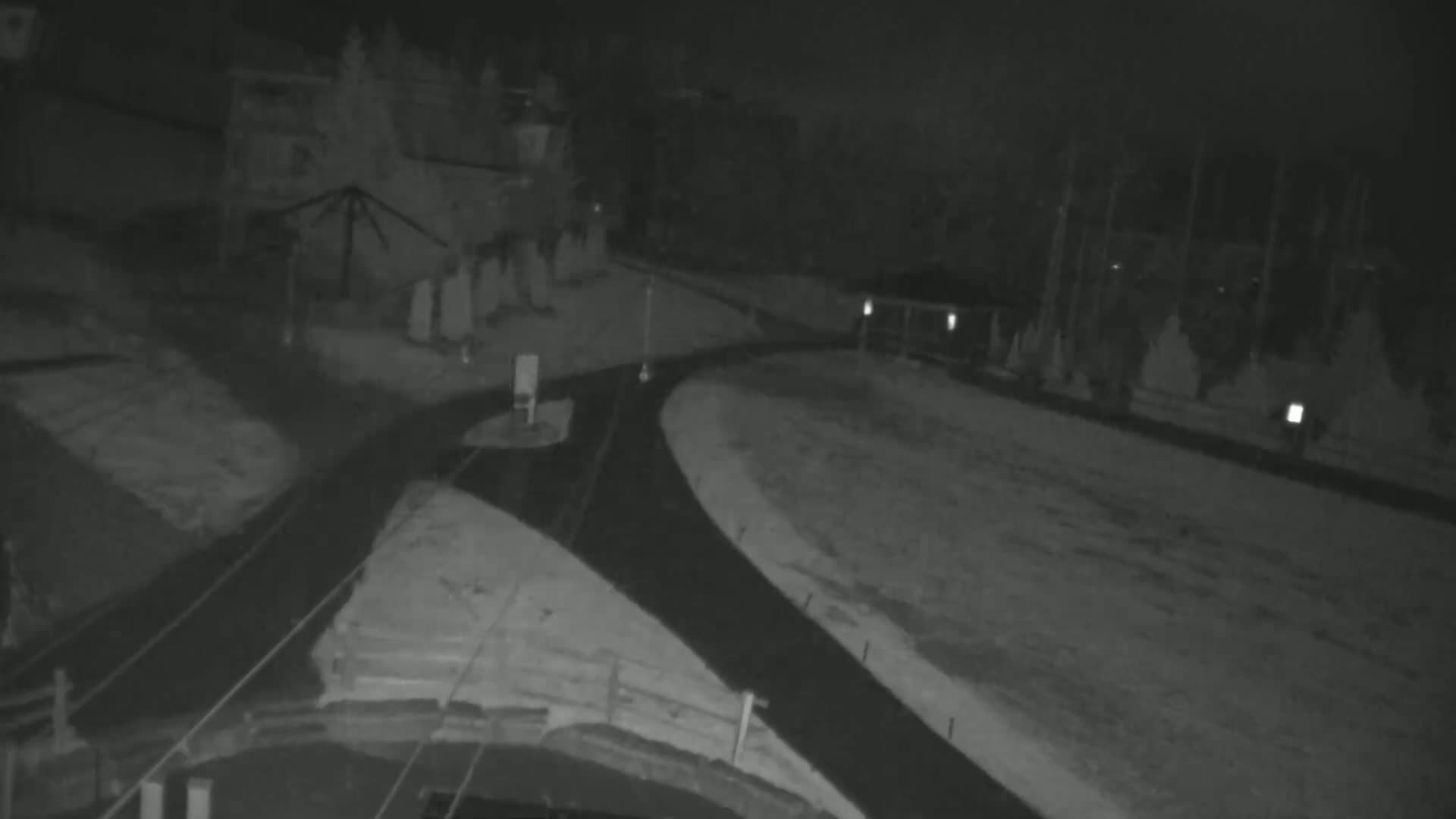 A nighttime, low-light view shows a snow-covered roadway curving to the right, with sparse vegetation and buildings visible in the background.